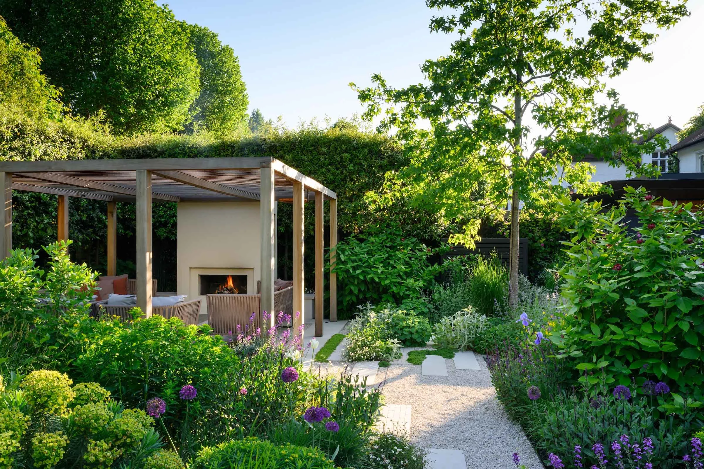 Timber pergola with fireplace and seating, surrounded by lush allium planting and a gravel path in a Putney garden by Kawauchi Rowe