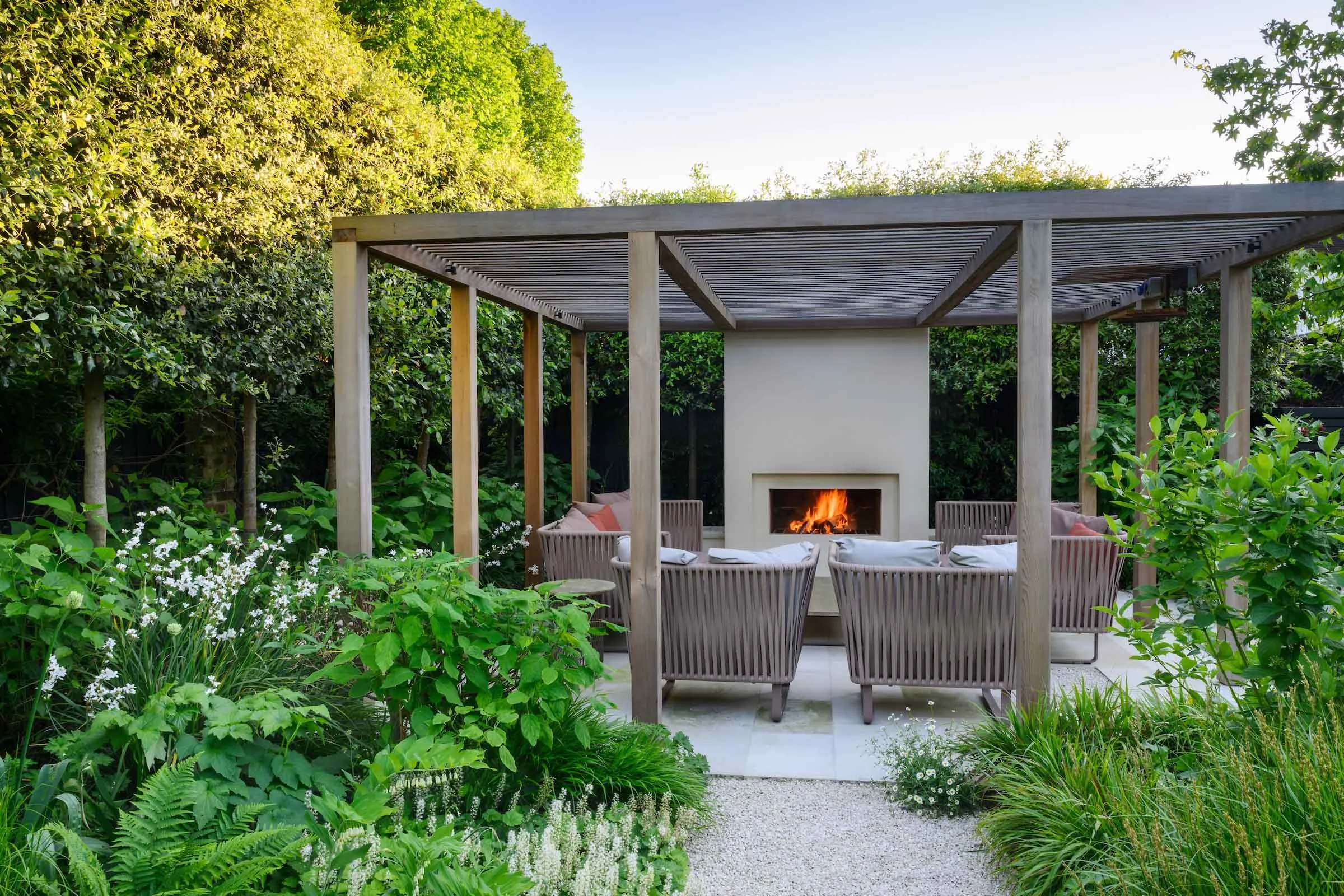 Bespoke timber pergola with a lit fireplace and dining seating among lush planting in a Putney garden by Kawauchi Rowe