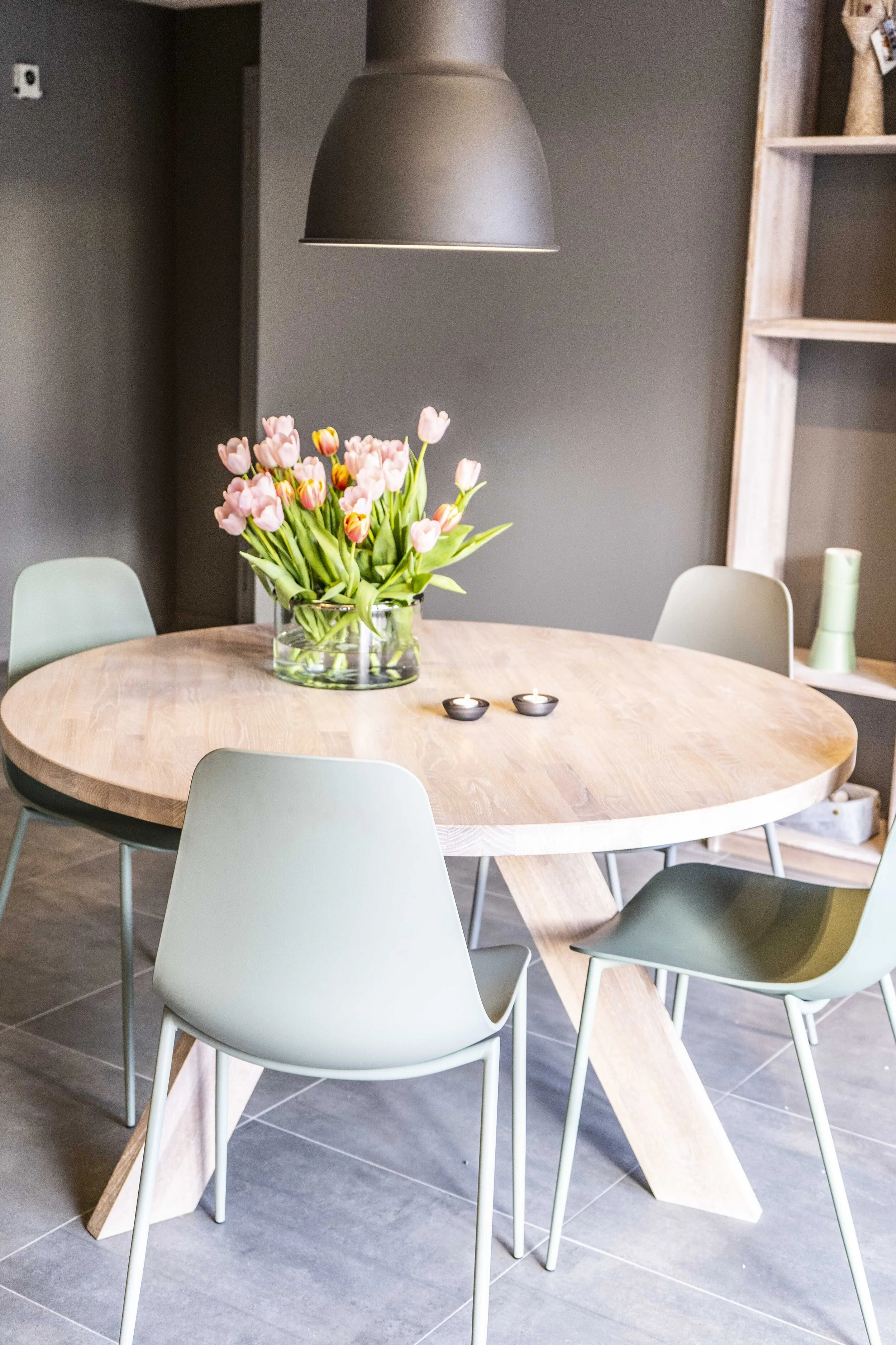Moderne spisestue med rundt bord av lyst tre, omgitt av fire grønne stoler. En vase med rosa tulipaner står på bordet ved siden av to telys. Grå lampeskjerm henger over bordet.
