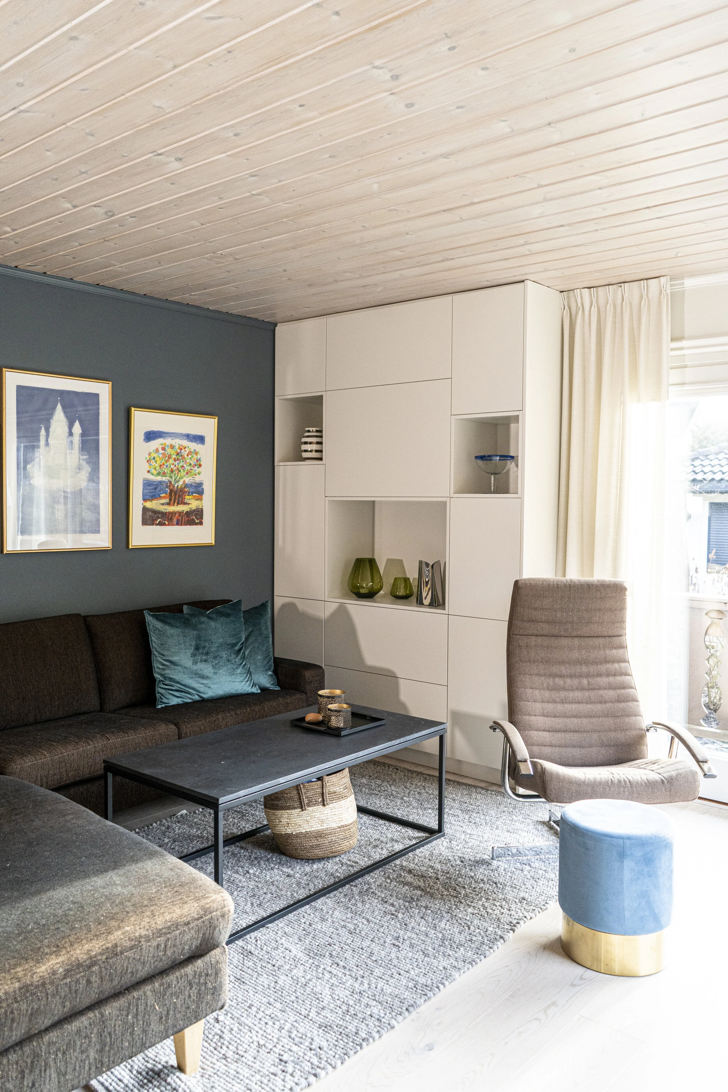 Moderne stue med mørk sofa, lyseblå pute og to bilder på veggen; en lenestol, et sofabord og et oppbevaringsmøbel med dekorasjoner; store vinduer med gardiner.