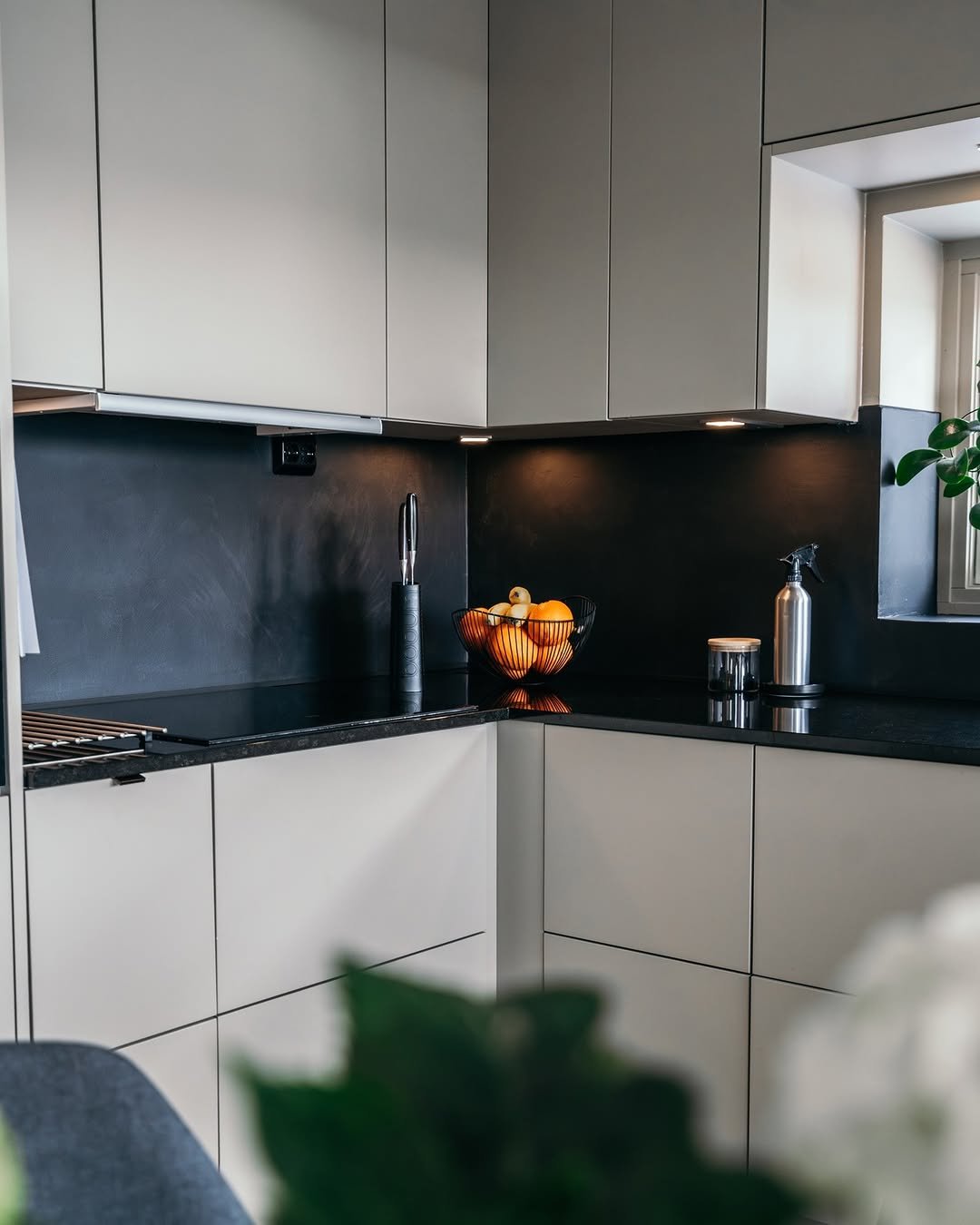 Moderne kjøkken med hvite skap, sort benkeplate, fruktkurv med appelsiner og andre frukter, og en sprayflaske.