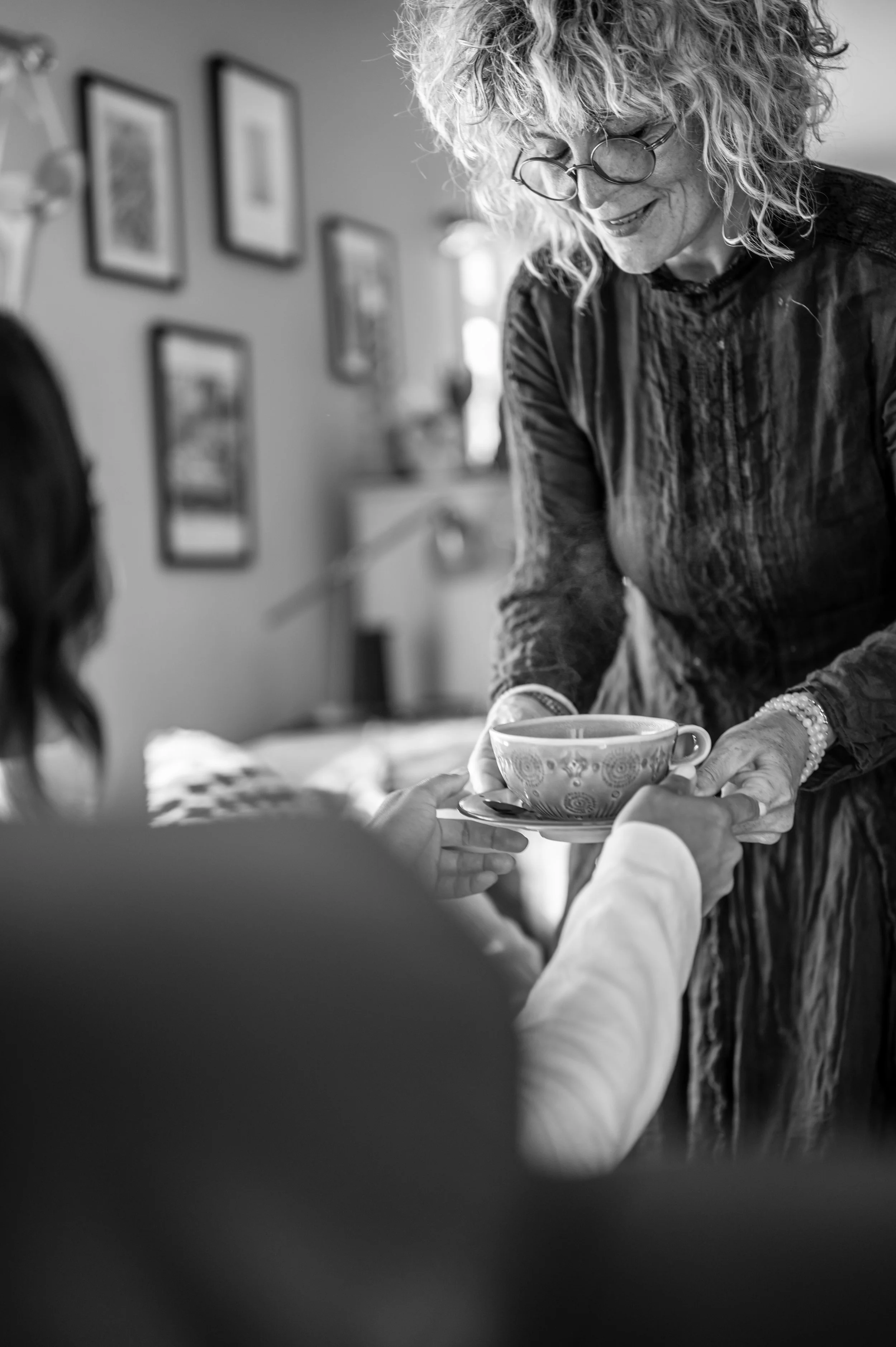 Woman with curly hair and glasses handing a teacup on a saucer to another person in a cozy home setting.
