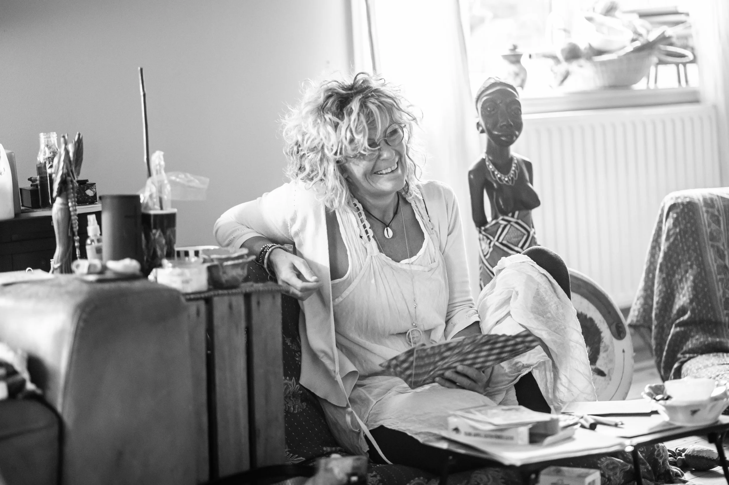 A woman with curly hair and glasses sitting on a sofa, smiling and looking at a photo or card in her hand. There is a woman with traditional African attire standing in the background near a radiator. The room has a cluttered table, and sunlight coming through a window.