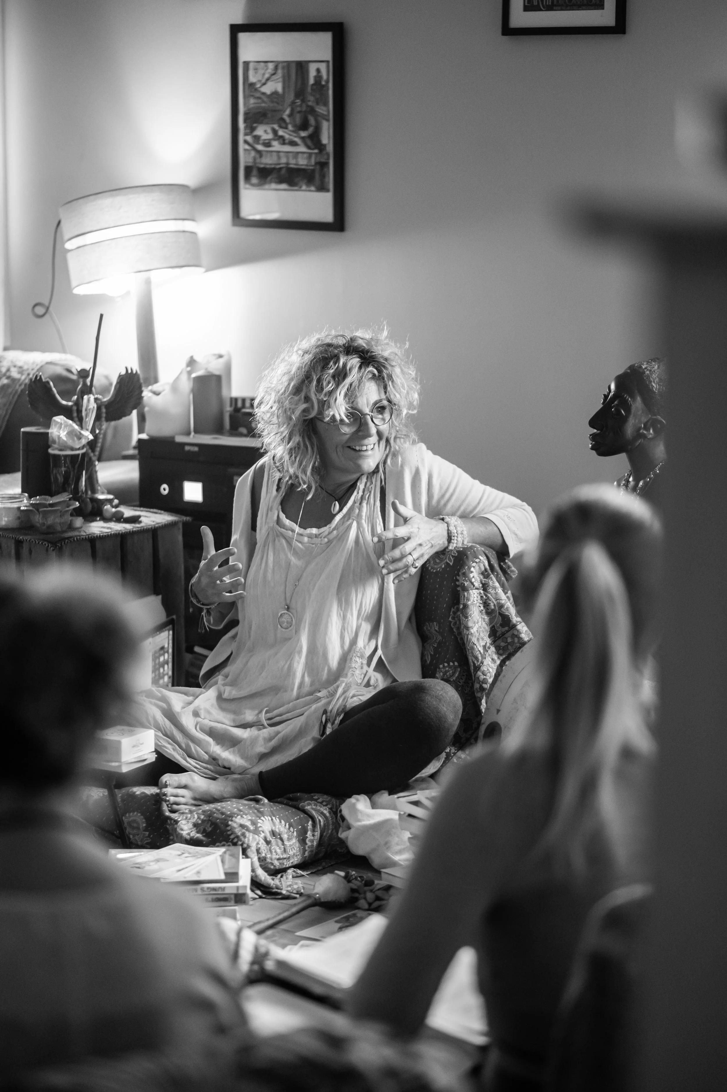 A woman with curly hair and glasses sitting cross-legged on a bed, smiling and talking to two people whose backs are to the camera in a cozy room with a lamp, framed pictures, and various decorative items.