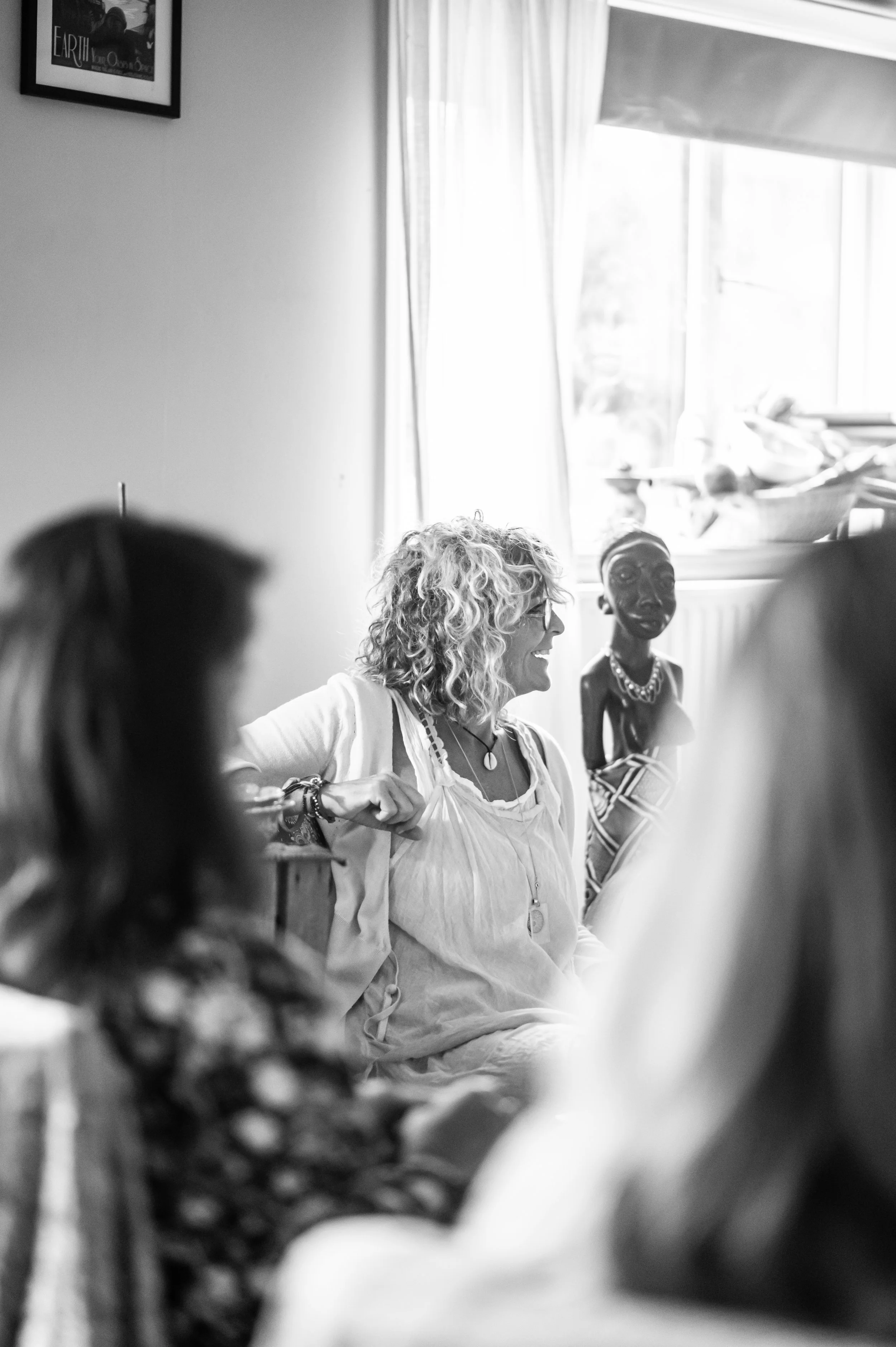 A group of women gathers in a bright room with large windows. One woman with curly hair is smiling and appears to be speaking. There is a colorful statue of a woman in the background near the window.