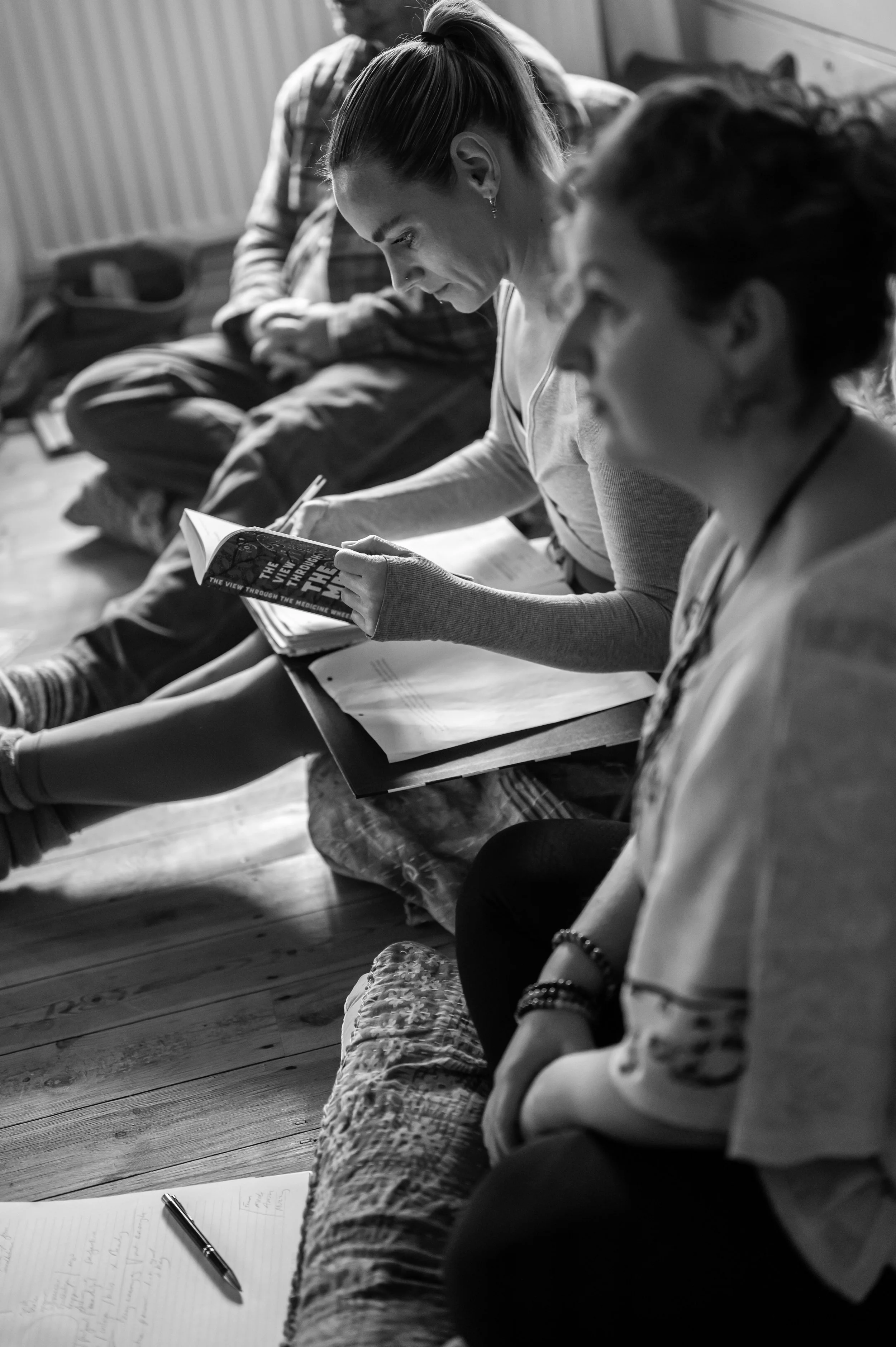 Four people sitting on the floor with notebooks and papers. One woman is reading a book, and others are engaged in discussion, in a casual indoor setting.