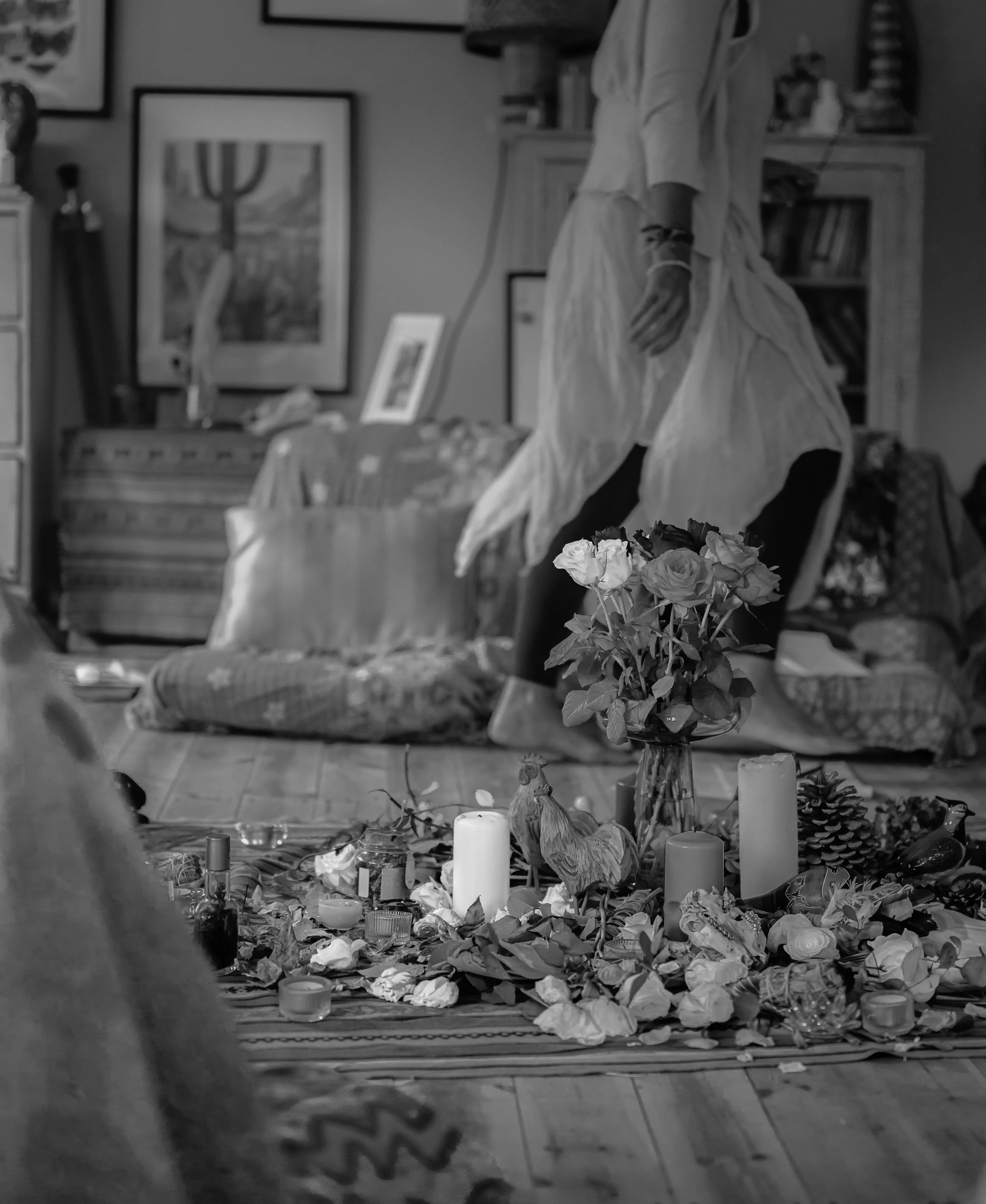 A woman in a light-colored dress kneels on a cushion behind a floral altar with candles, flowers, and decorative objects on a rug in a living room with framed artwork and bookshelves.