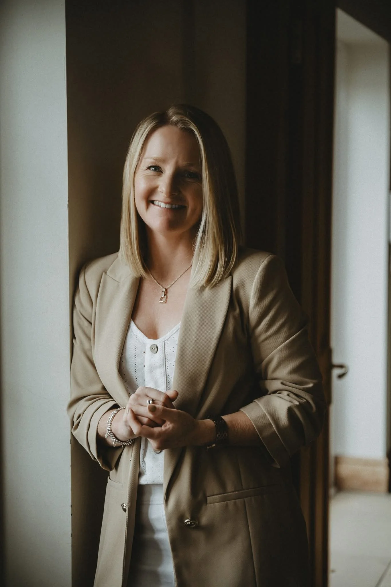 A woman with shoulder-length blonde hair, wearing a beige blazer over a white top, smiling and standing in a doorway.