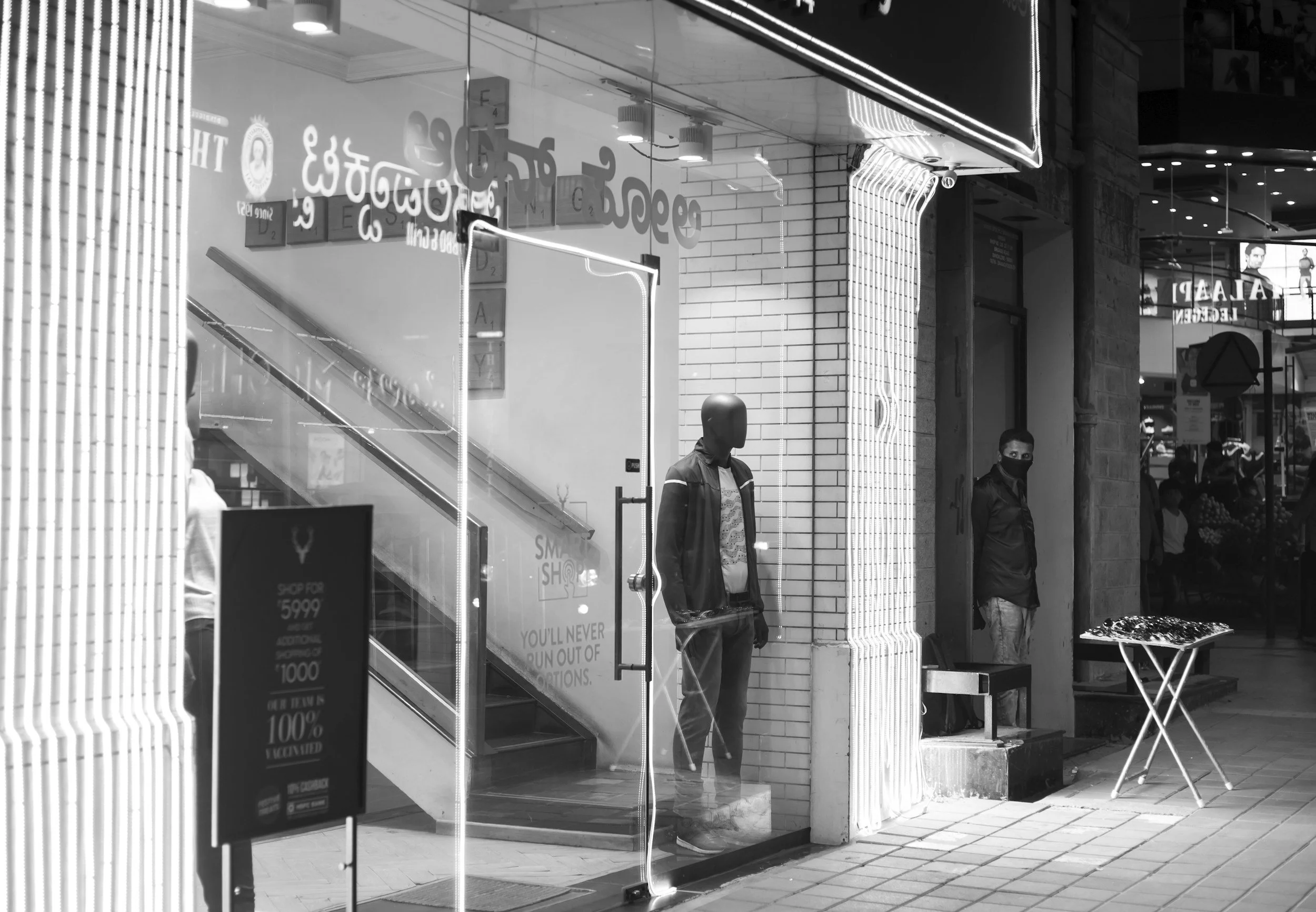 A storefront with glass doors and windows at night, featuring modern neon lighting and advertising signs. two men with face masks stand outside, and there is a street vendor with a table displaying goods nearby.