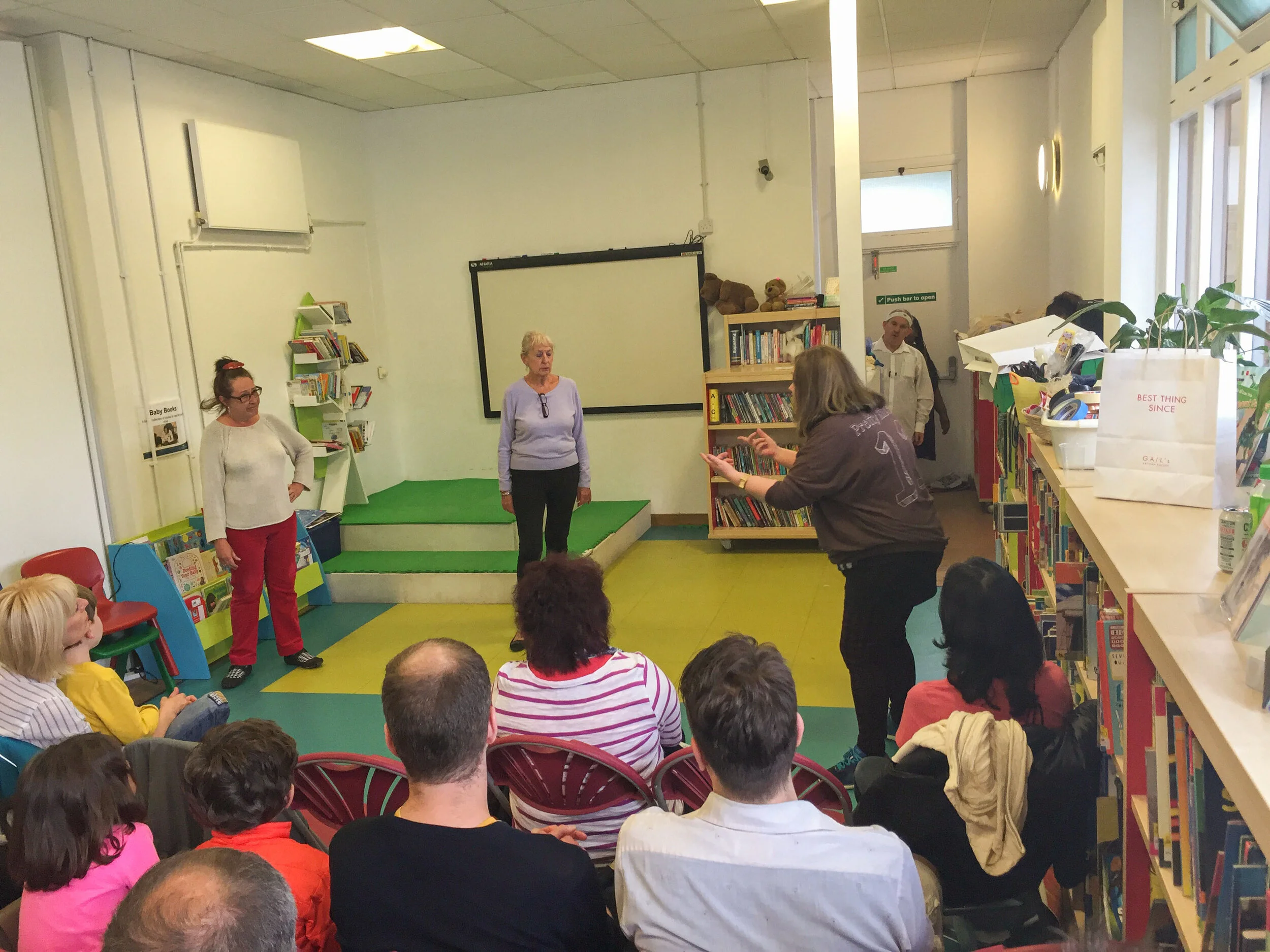A group of children and adults in a library, with a woman in black gesturing and talking to a woman in purple who is standing on a small green stage, as others watch, during a presentation or storytime session.