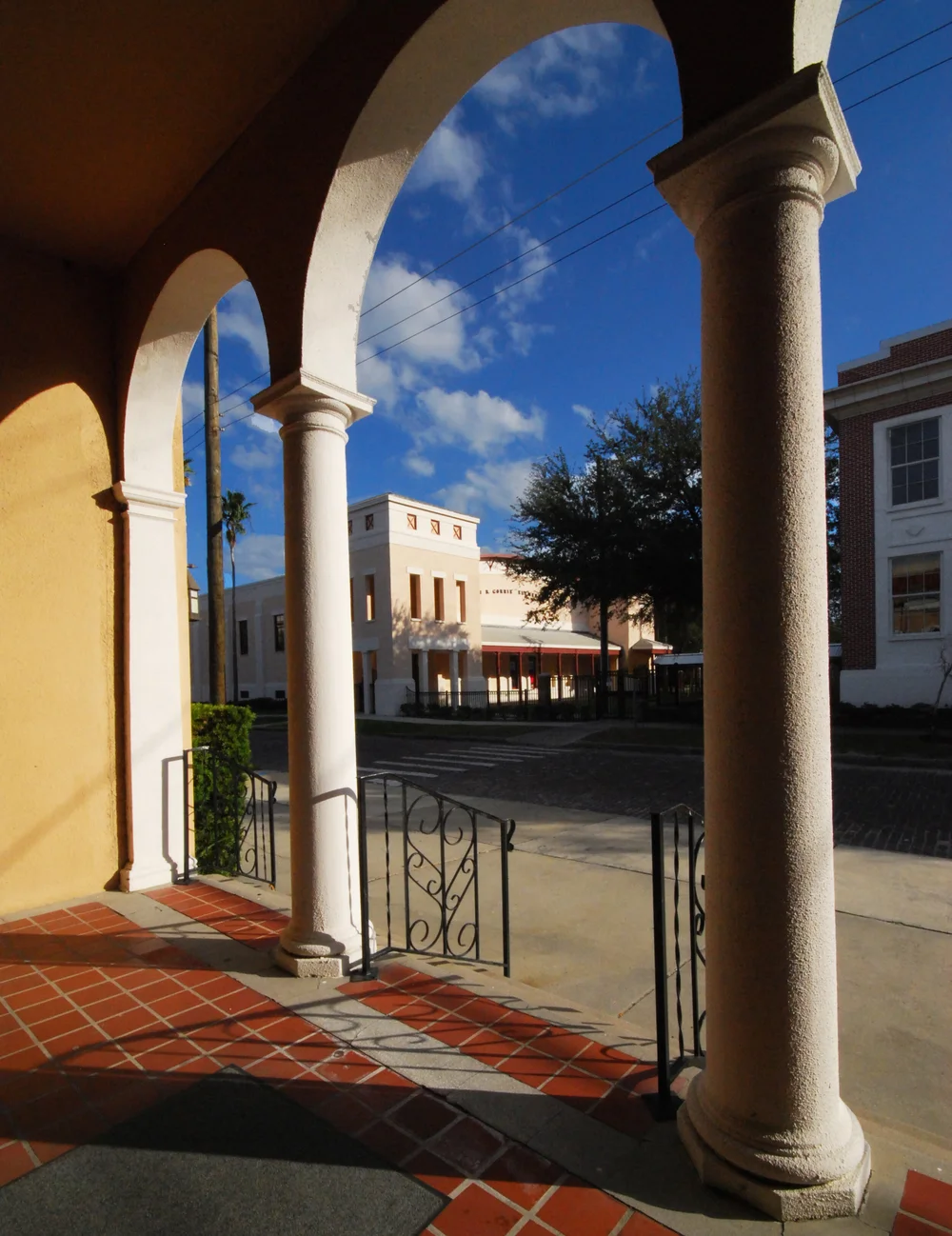 Portrait-format color architectural photograph made from within or immediately at FMM's entrance arcade. The ochre wall, three white arches and columns, terracotta tiled floor and wrought-iron gates occupy the foreground and frame the completed John B. Gorrie Elementary coliseum and part of the older brick school across the paved way. The portfolio's filename establishes the stated vantage point; the page does not identify the photographer, capture date, alterations visible in FMM fabric or any formal relationship between FMM and the school project.