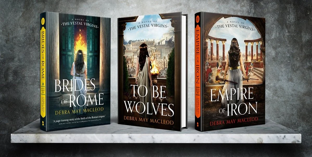Three books from the 'Vestal Virgins' series by Debra May Macleod displayed on a marble surface against a gray wall. The first book is titled 'Brides of Rome,' featuring a woman in white in front of an ancient Roman door with fire behind it. The second book is 'To Be Wolves,' showing a woman in ancient Roman attire overlooking a cityscape with a fire burning in the distance. The third book is 'Empire of Iron,' with a woman in Roman-style clothing standing in front of an ancient Roman architectural backdrop with columns and arches.
