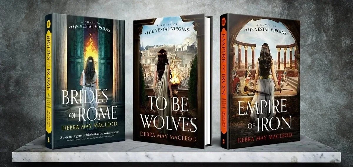 Three books from the Vestal Virgins series by Debra May MacLeod displayed on a marble surface against a gray wall. The titles are 'Brides of Rome,' 'To Be Wolves,' and 'Empire of Iron,' each with cover art depicting ancient Roman themes and female characters.