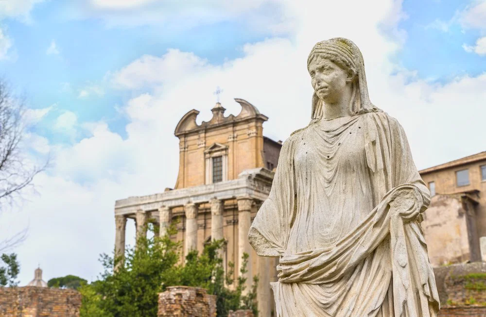 A marble statue of a woman in front of an ancient building, under a partly cloudy sky.