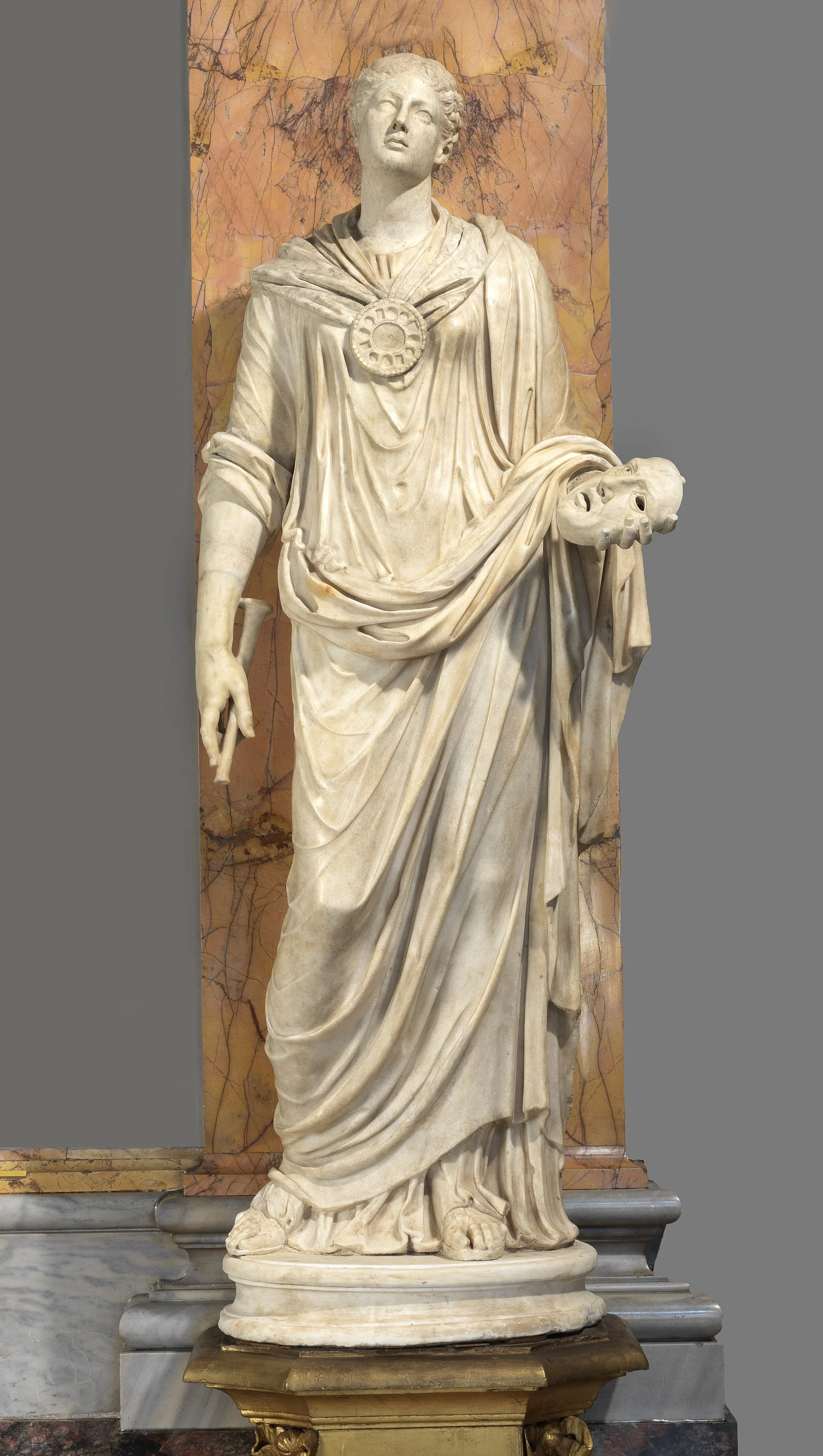 Marble statue of a woman with a saddened expression, holding a severed head in her left hand, with draped clothing and a circular emblem on her chest, set against a marble background.