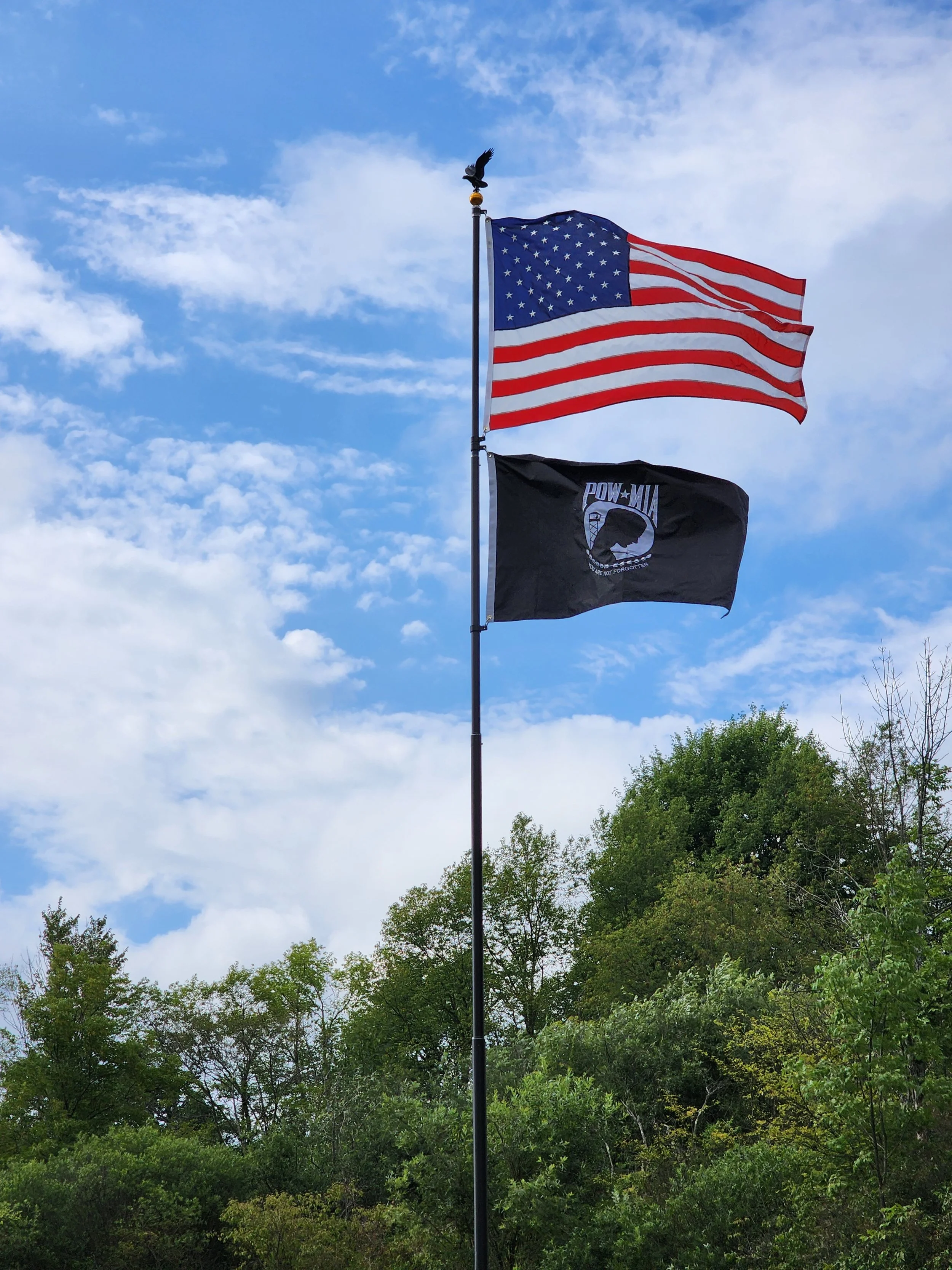 Three flags flying on a pole against a partly cloudy sky, with green trees in the background. The top flag is the American flag, the middle flag is black with a POW/MIA emblem, and the bottom flag is plain black.