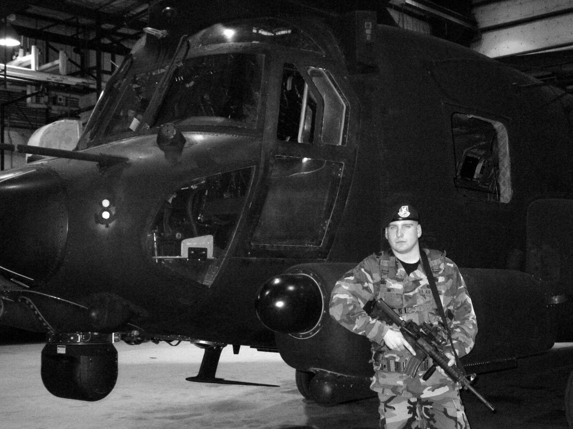 A soldier in camouflage uniform holding a rifle standing in front of a military helicopter inside a hangar.