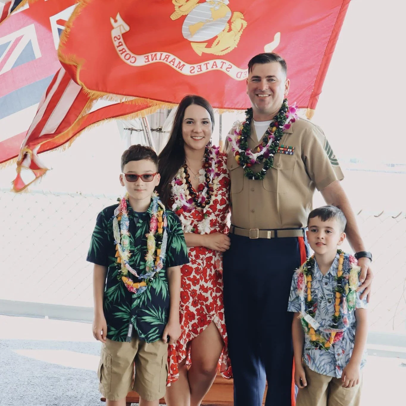 Group of people at a celebration, including a woman in a red floral dress, a man in a military uniform, and two boys in tropical shirts, all wearing lei necklaces, standing under a large red United States Marine Corps flag.