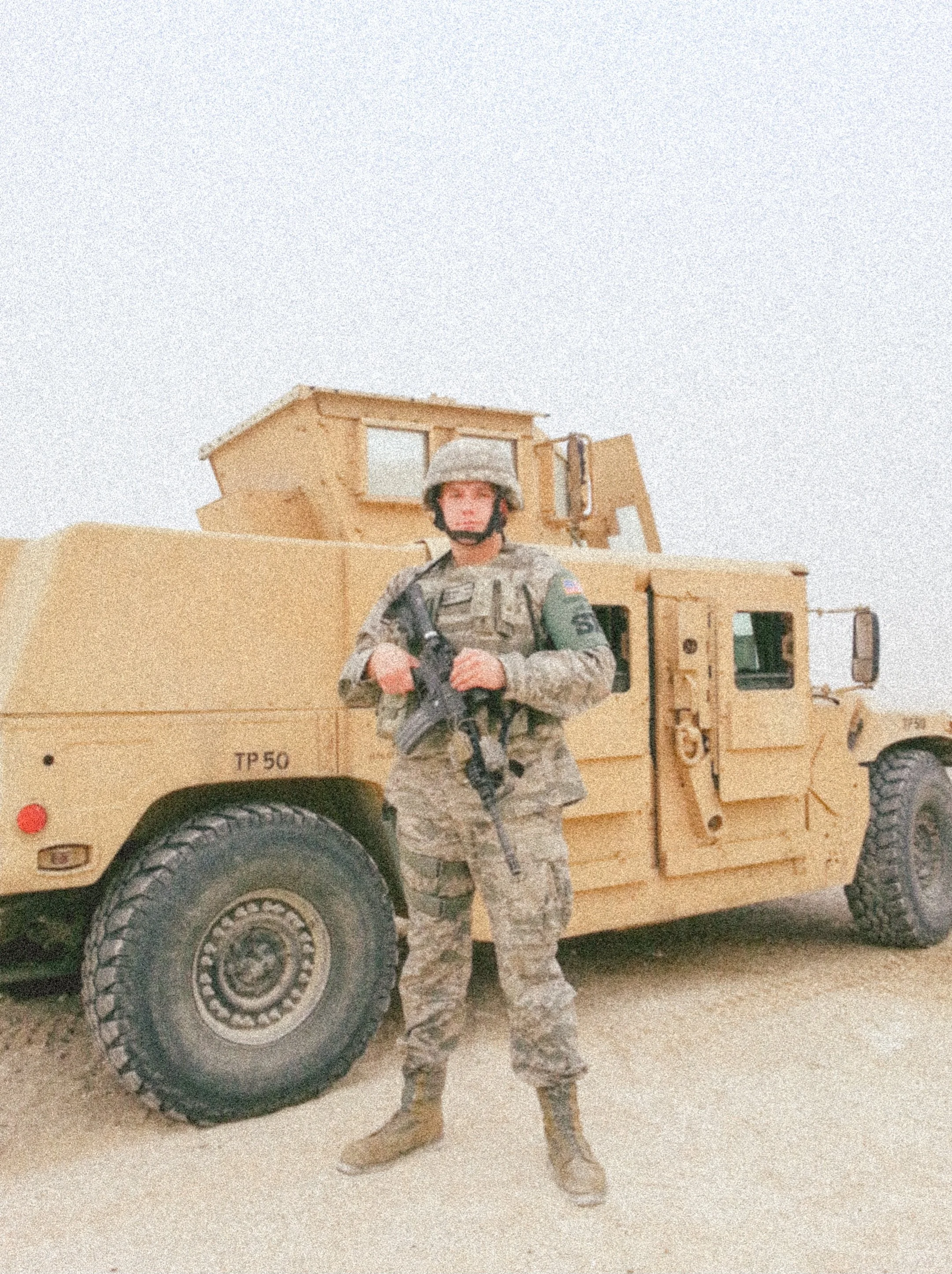 A soldier in uniform stands in front of a large military vehicle, holding a rifle, with a desert landscape and a clear sky in the background.
