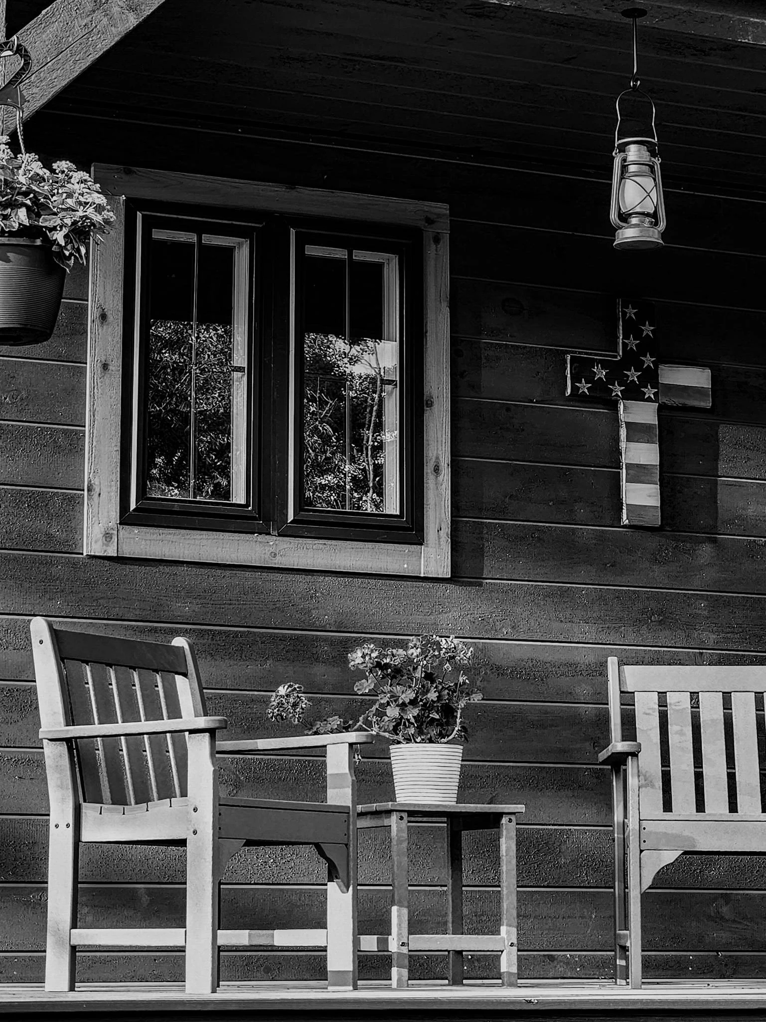 Porch with two wooden chairs and a small table with a potted plant, window with wooden frame, hanging lantern, and a star-shaped American flag on the dark wooden wall.