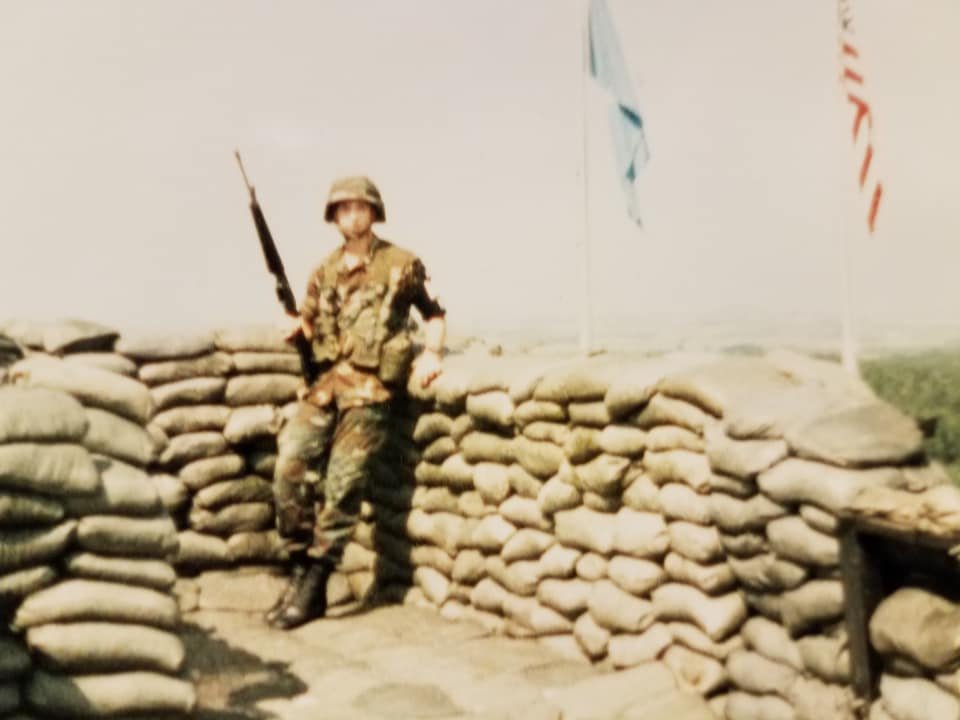 A soldier in camouflage uniform standing by a sandbag wall at a military outpost, holding a rifle, with flags flying in the background.