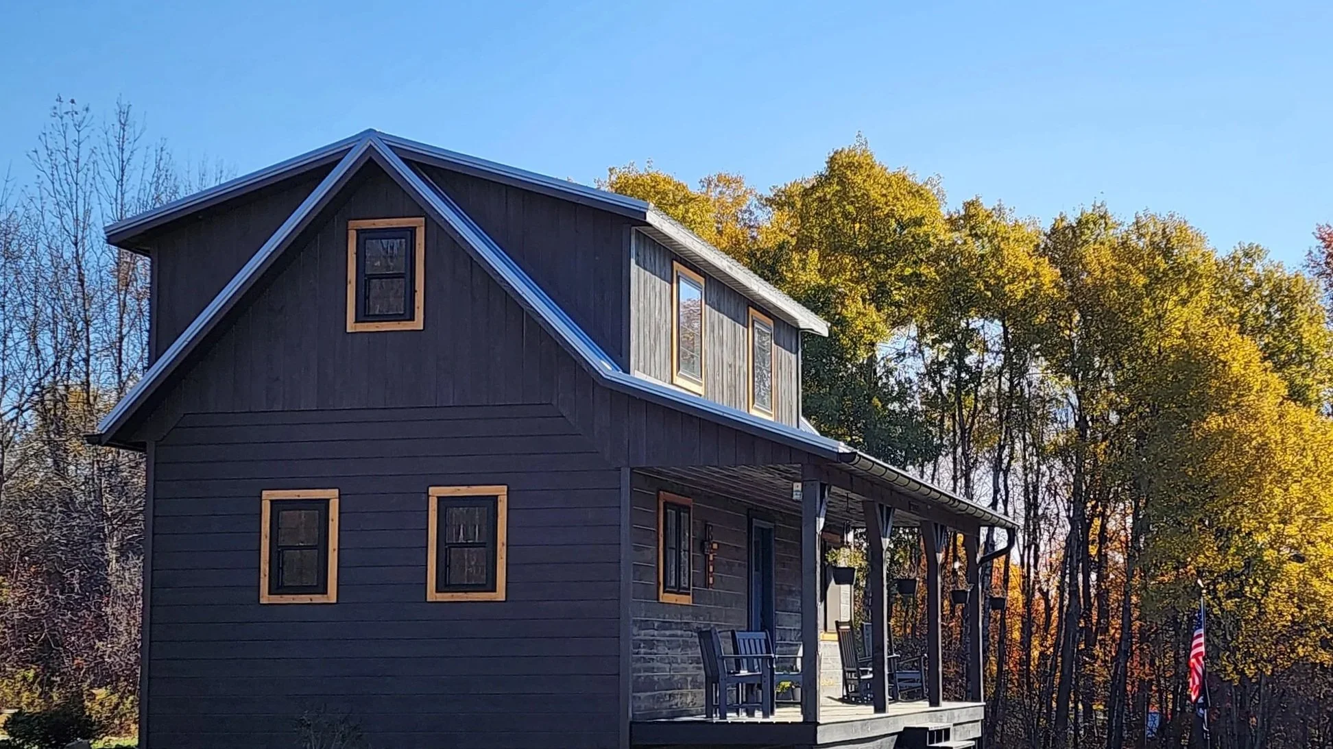 A dark gray house with three levels, surrounded by fall-colored trees; the house has several windows with wooden frames and a covered porch.