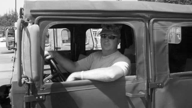 A young man wearing sunglasses and a t-shirt sitting in the driver’s seat of a vintage or classic vehicle, possibly a Jeep or similar, with the window rolled down.