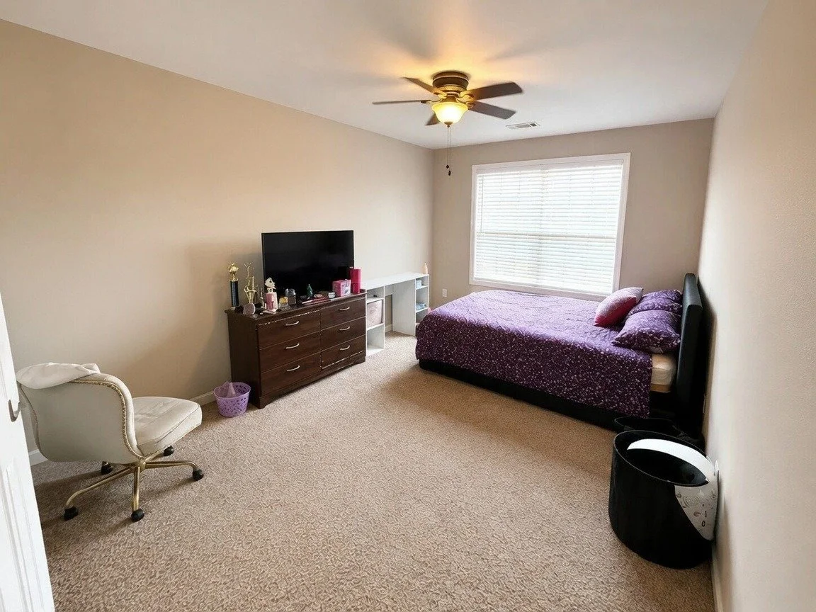 A bedroom with a bed dressed in purple bedding, a wooden dresser with trophies, a white chair, a mirror leaning against the wall, a TV, and a window with blinds. There are framed certificates on the wall and various items on the dresser.