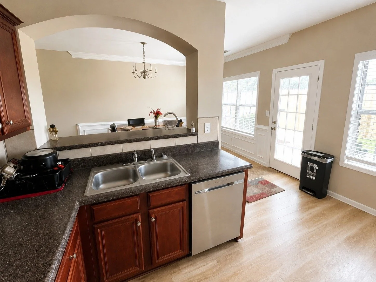 Kitchen with a double sink, dark granite countertops, wooden cabinets, and a view into a dining area with a chandelier, dining table, and chairs. There's a door with glass panels leading outside, and windows with blinds letting in natural light. A tr
