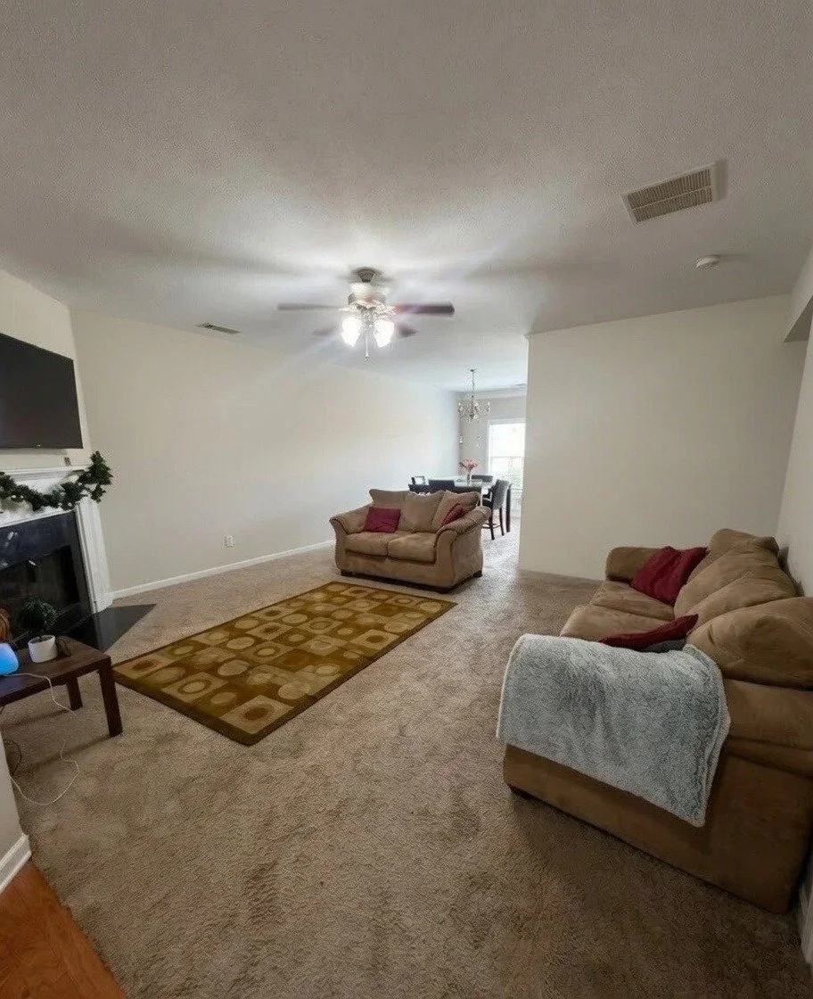 A cozy living room with beige sofas, a fireplace decorated with greenery, framed photos on a coffee table, a patterned rug, and a dining area in the background with a chandelier and large window.