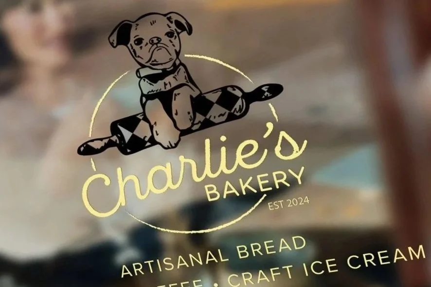 Logo of Charlie's Bakery featuring a cute puppy sitting on a rolling pin with the text "Charlie's Bakery" and additional text about artisanal bread, coffee, and ice cream.