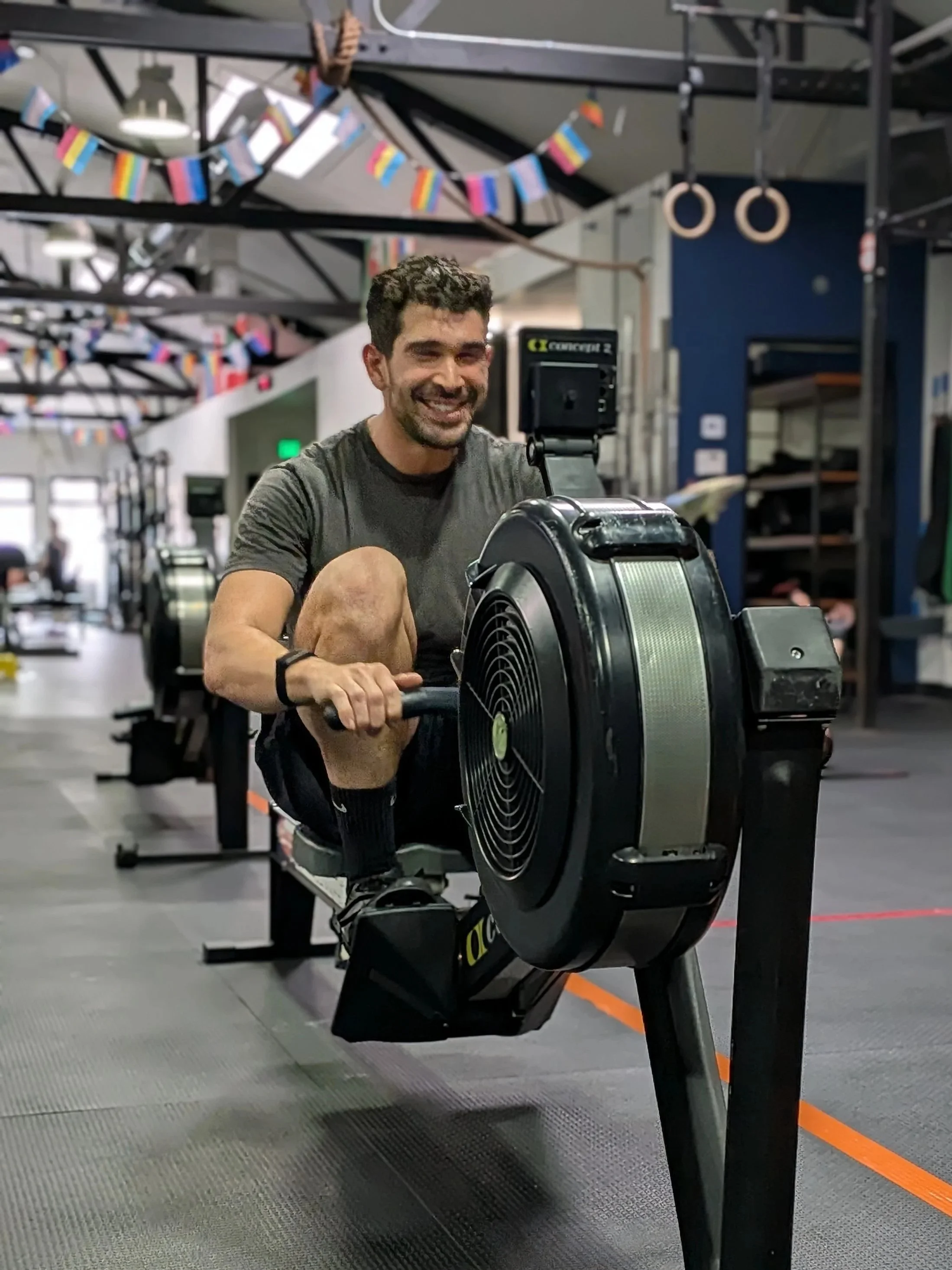 Man smiling while using a rowing machine in a gym decorated with colorful flags and gym equipment in the background.