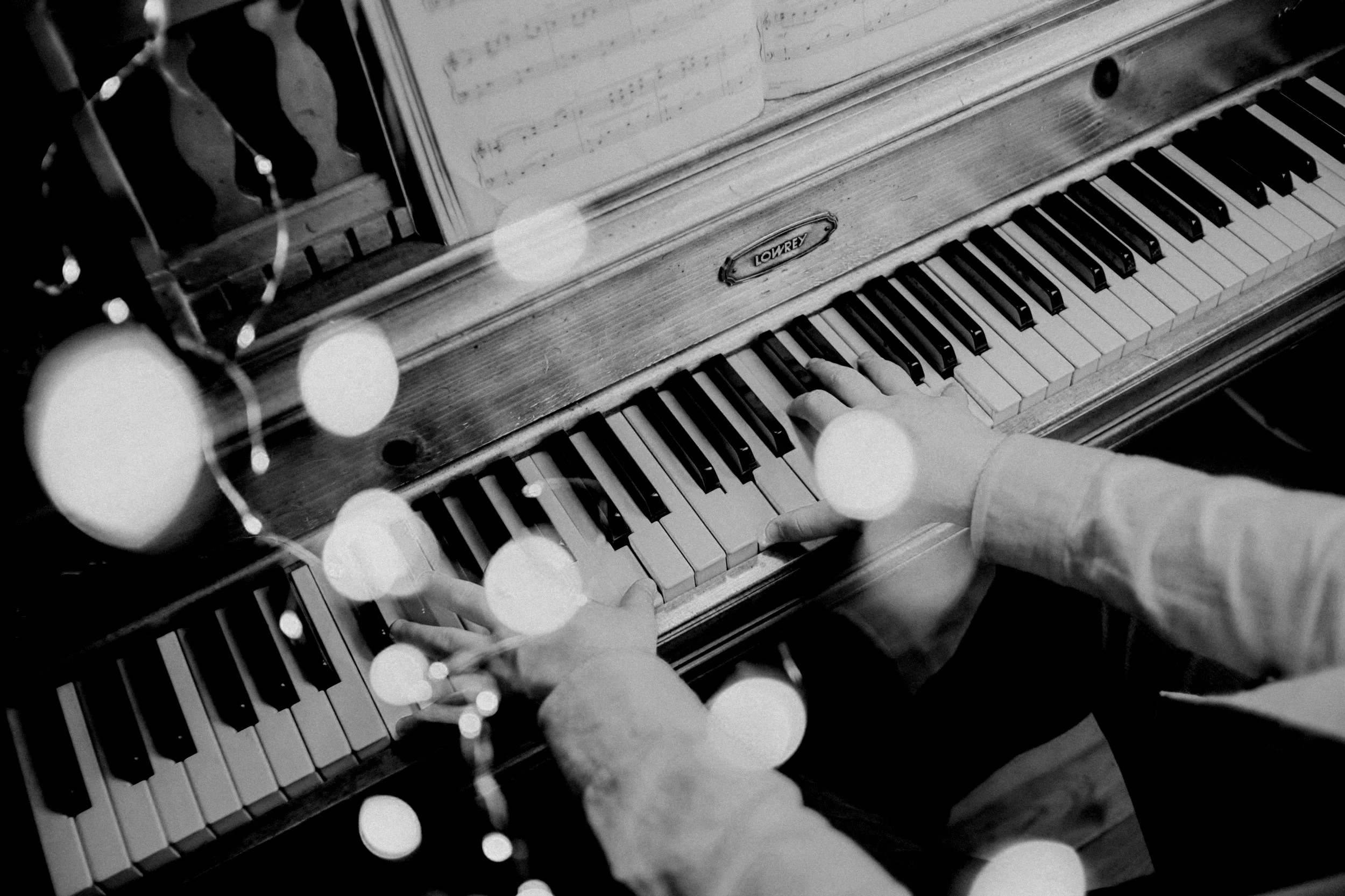 Black and white photo of a person playing a vintage lowrey piano, with sheet music on top, and string lights out of focus in the foreground.