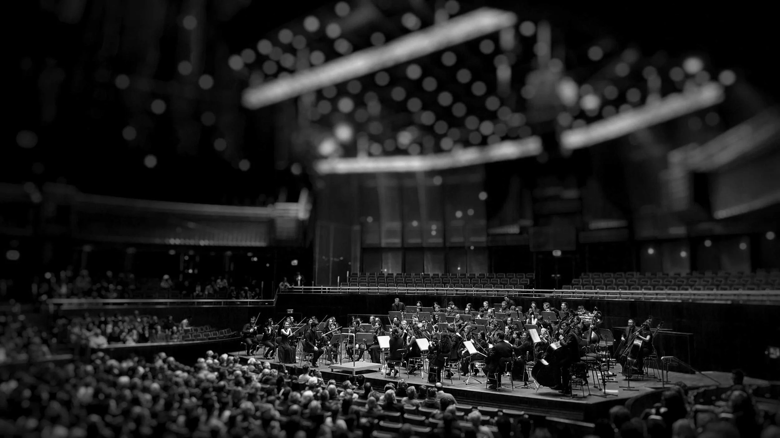 A large orchestra performing on stage inside a concert hall, with an audience watching.