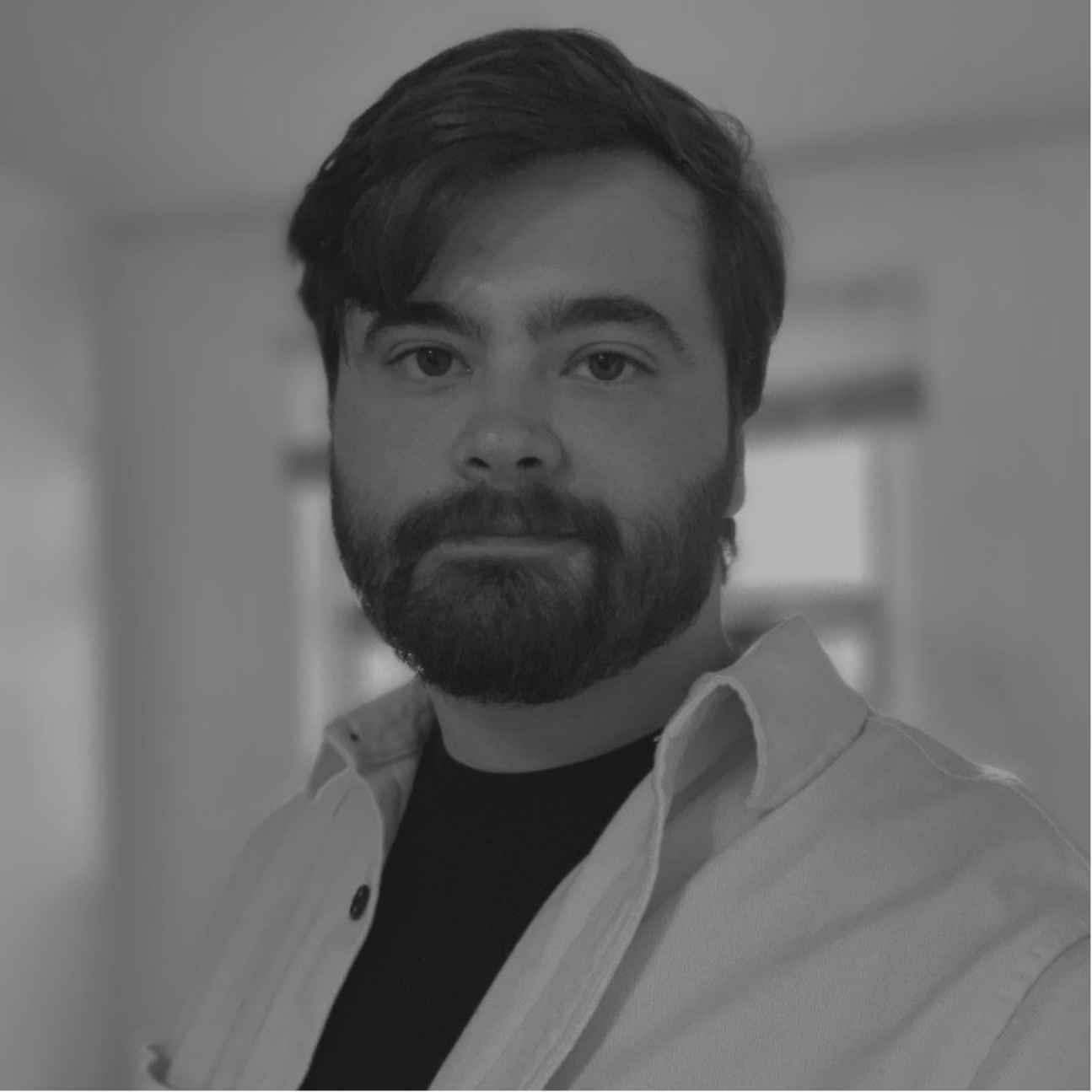 Black and white photo of a man with dark hair and a beard, wearing a collared shirt and a t-shirt, looking at the camera with a serious expression.