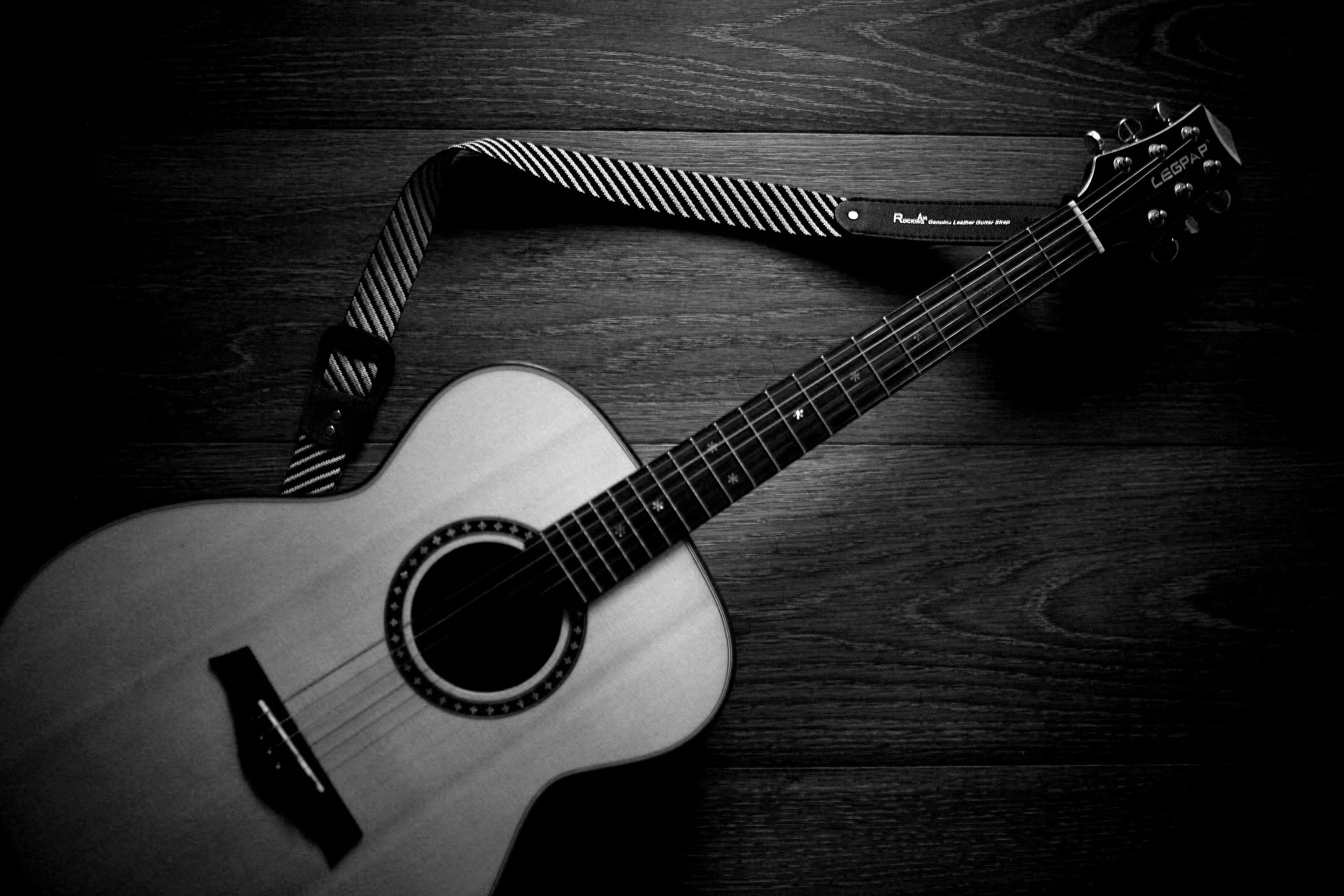 A black and white photo of an acoustic guitar lying on a wooden floor, with the guitar's neck angled toward the top right corner of the image.