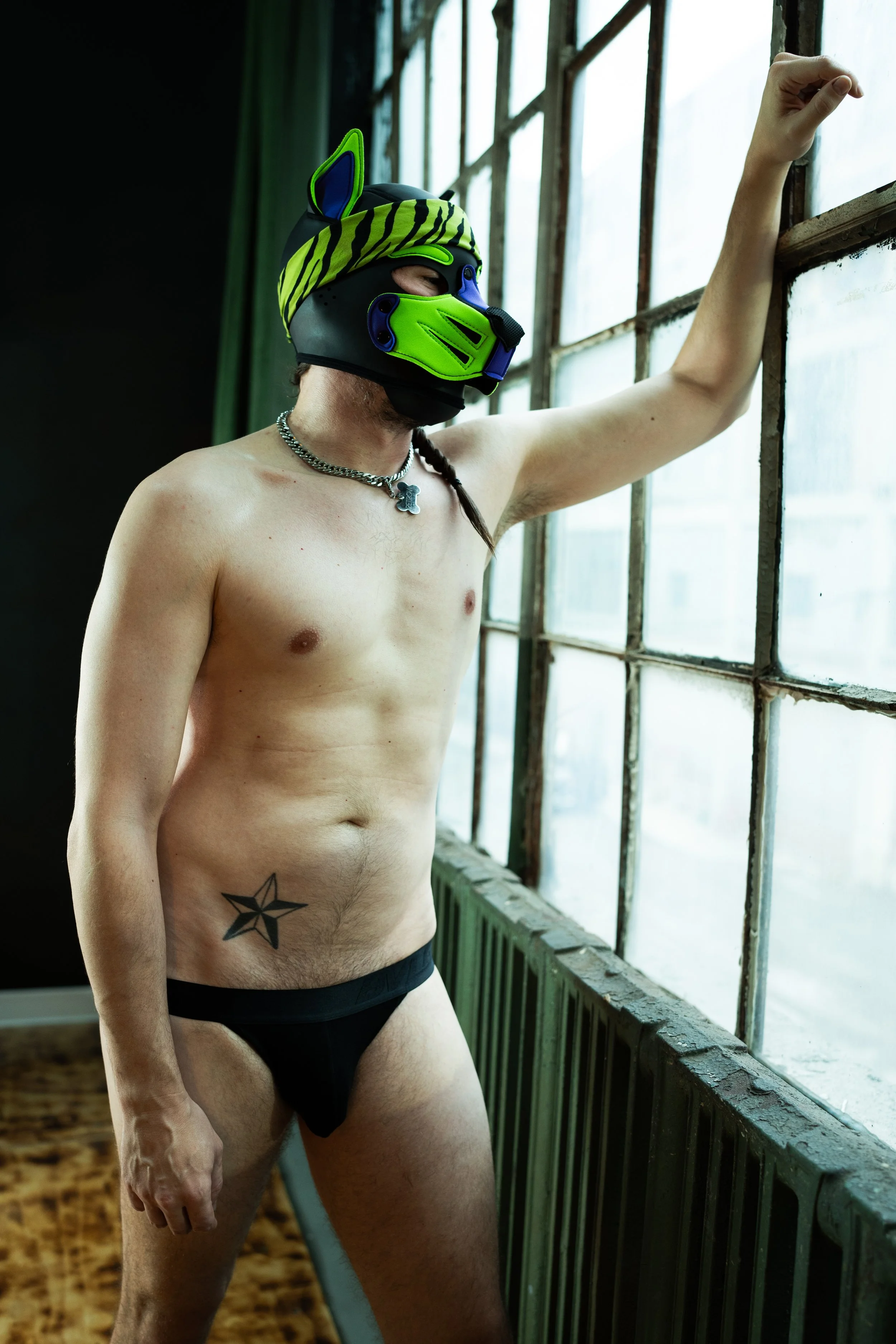 A shirtless man wearing a colorful dog mask, a striped headband, a silver chain necklace with a dog tag, and black underwear, standing by a large window in an industrial-style room.