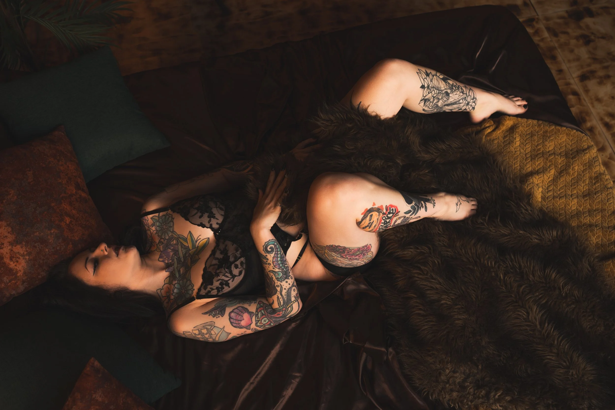Two tattooed women lying on a bed with dark sheets and pillows, one with black hair and the other with brown hair, surrounded by pillows and a furry blanket.