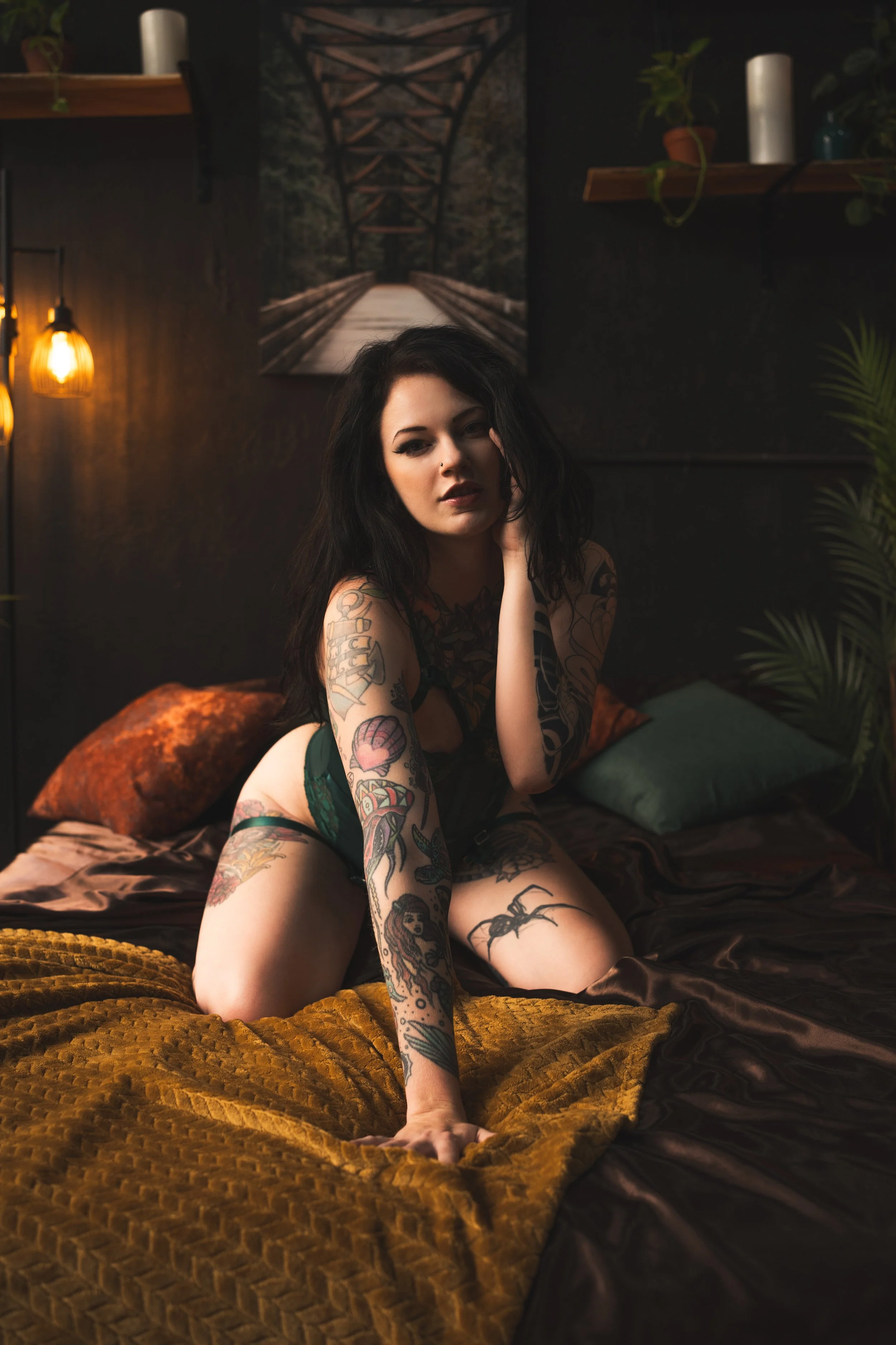 A woman with tattoos and dark hair sitting on a bed, in a dimly lit room with dark walls, velvet bedding, and warm lighting, with artwork of a bridge on the wall behind her.