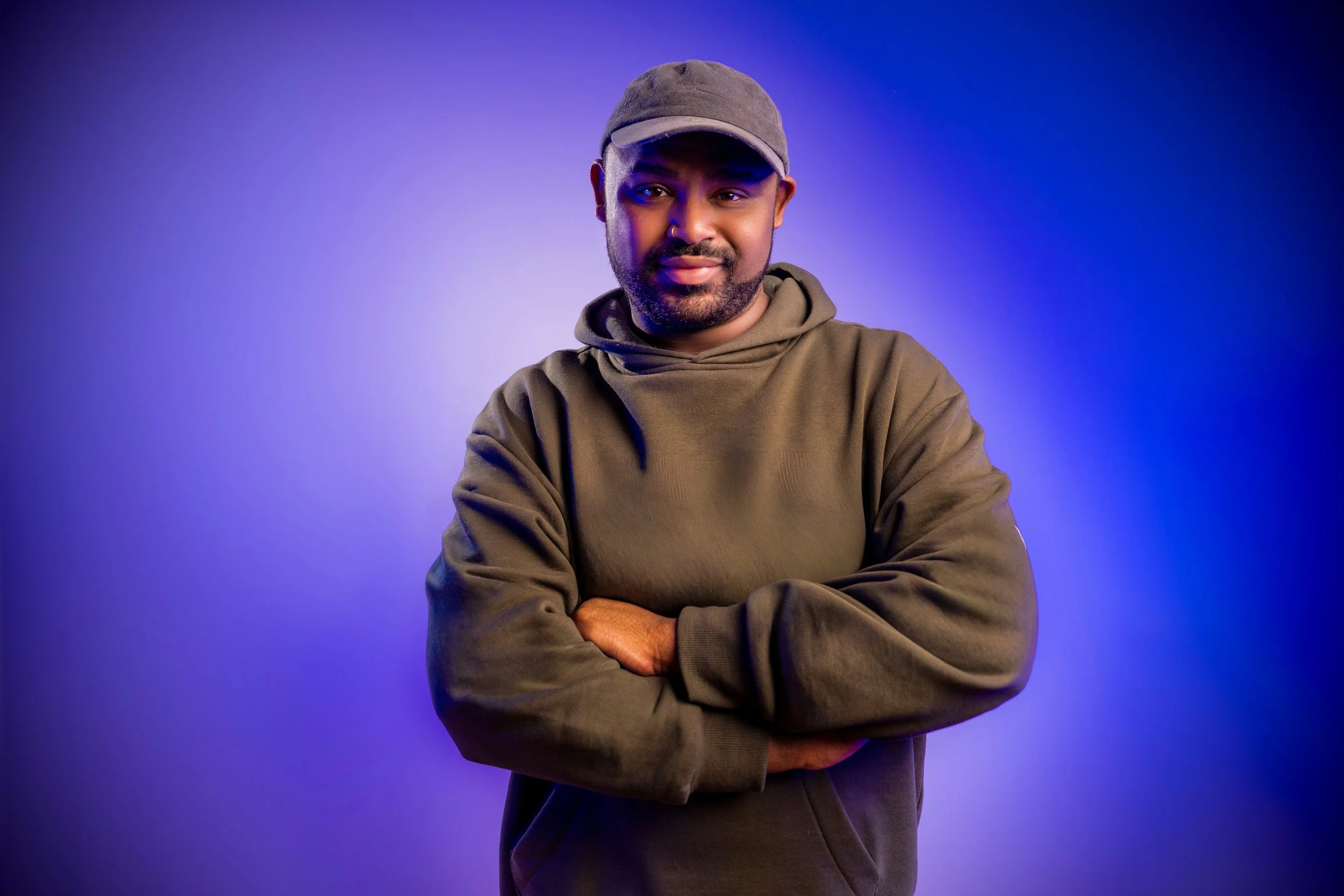 Portrait of a man wearing a gray cap, gray hoodie, standing against a purple and blue gradient background with arms crossed.