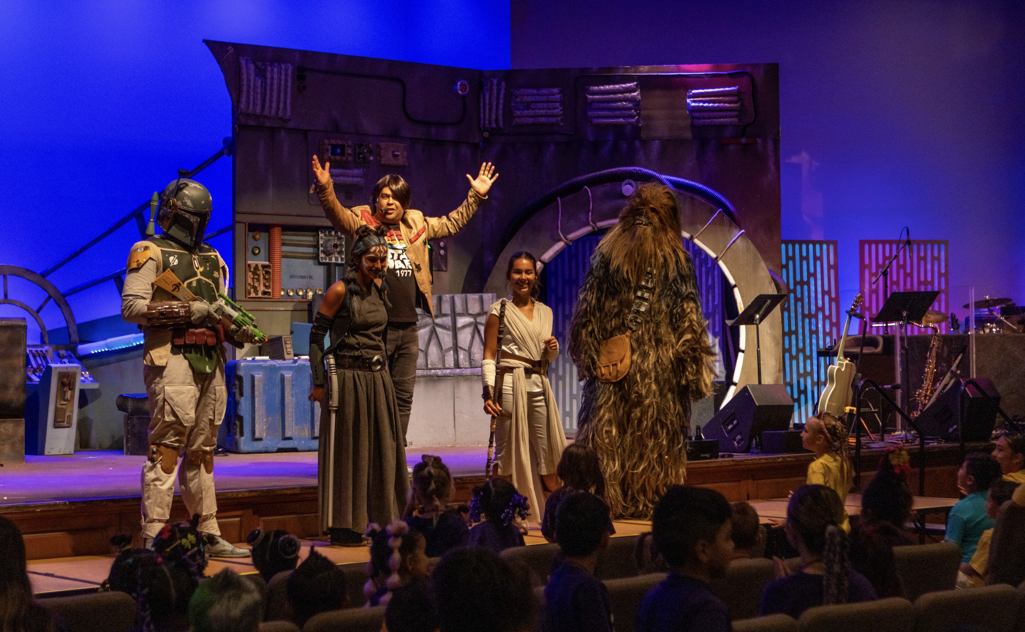 Children and a young woman with face paint playing a clapping game indoors with wooden decor and a large digital clock showing 11:39 in the background.