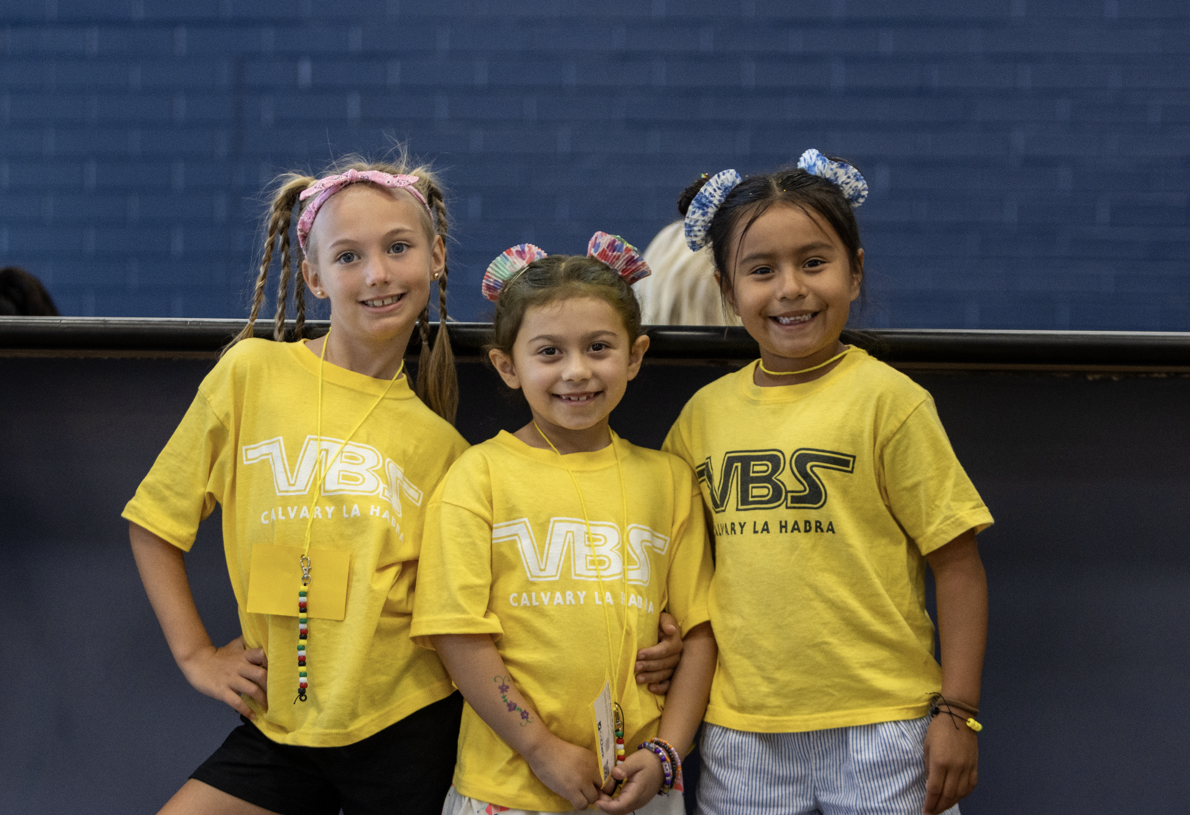 Two young boys wearing yellow t-shirts with 'SUMMER' written on them, standing close together with their arms around each other, smiling in a large room with beige chairs and other children in the background.