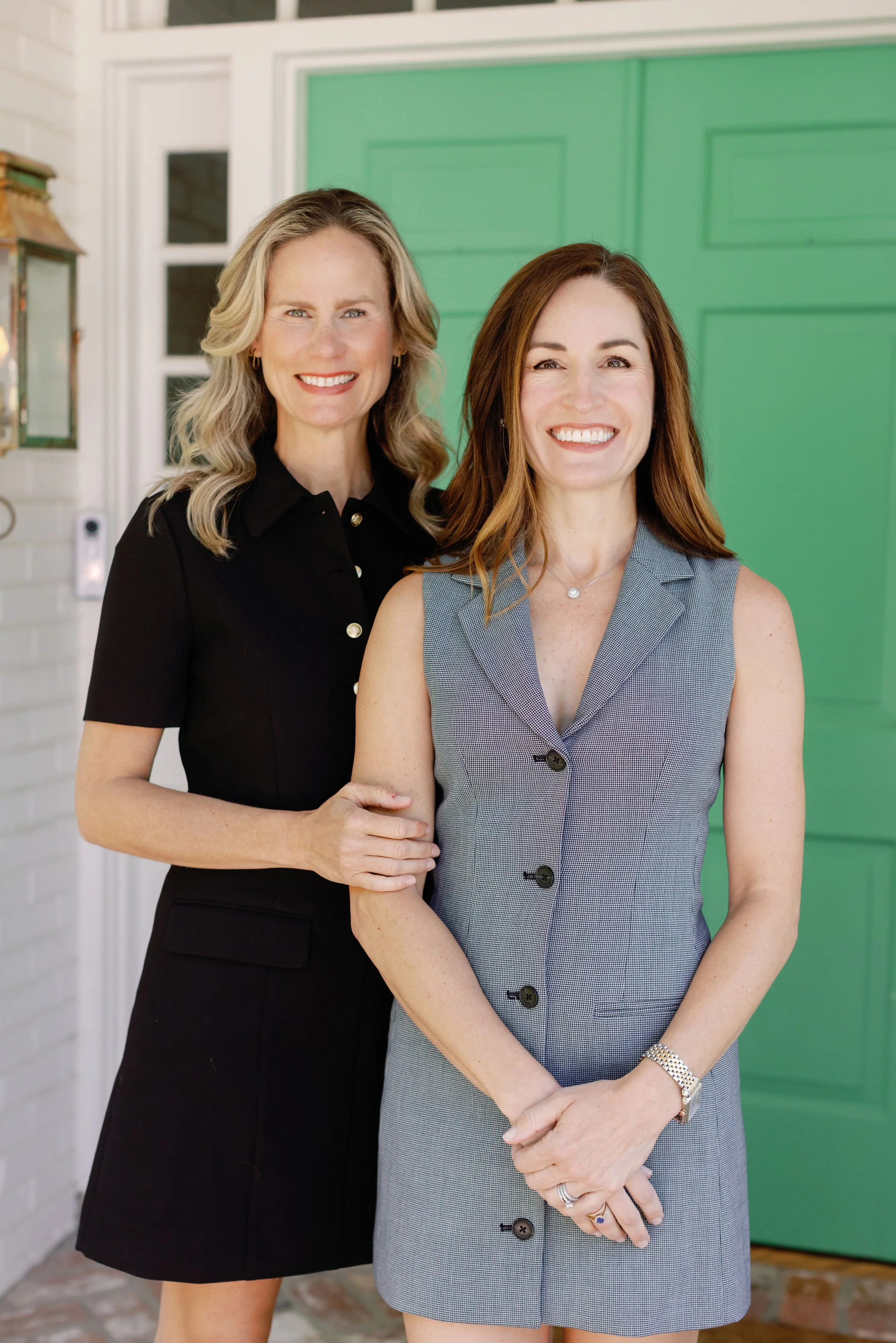 Two women standing close together smiling outdoors in front of a green door, with a background including white siding and stairs.