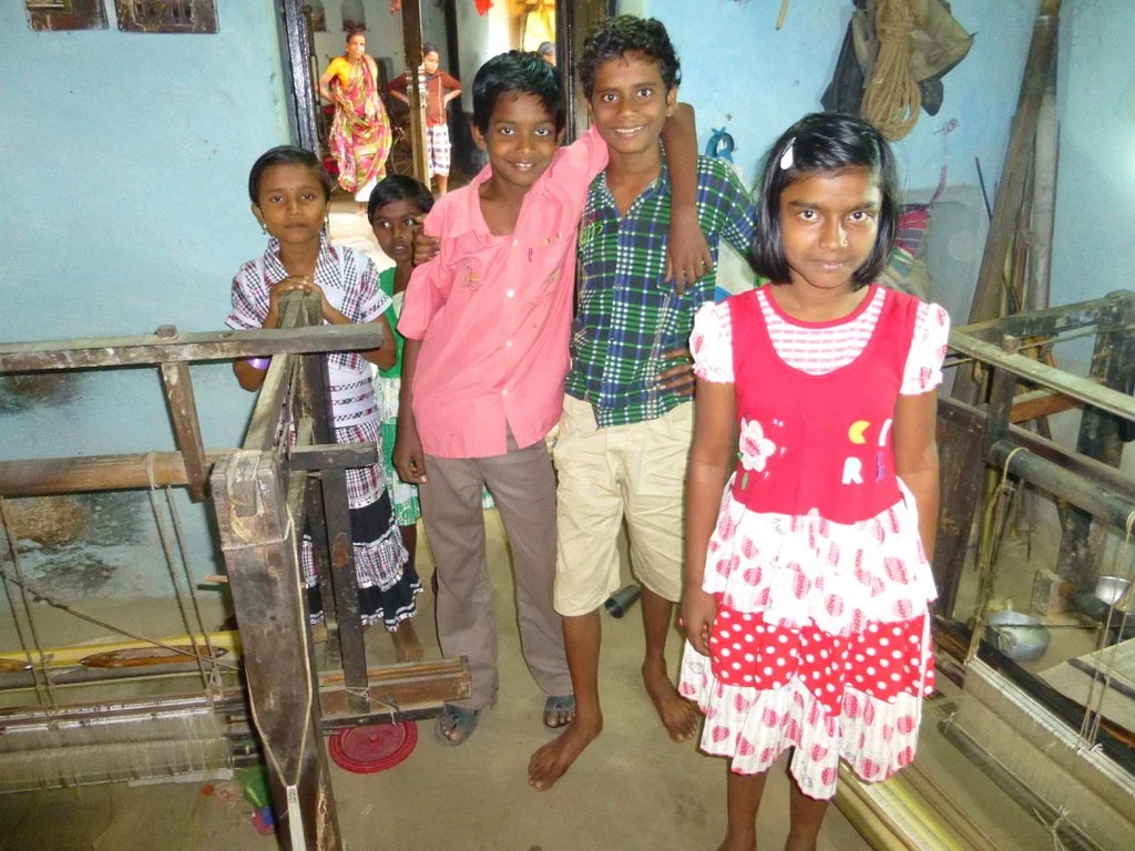 Group of six children inside a room with traditional weaving tools and colorful walls, smiling at the camera.