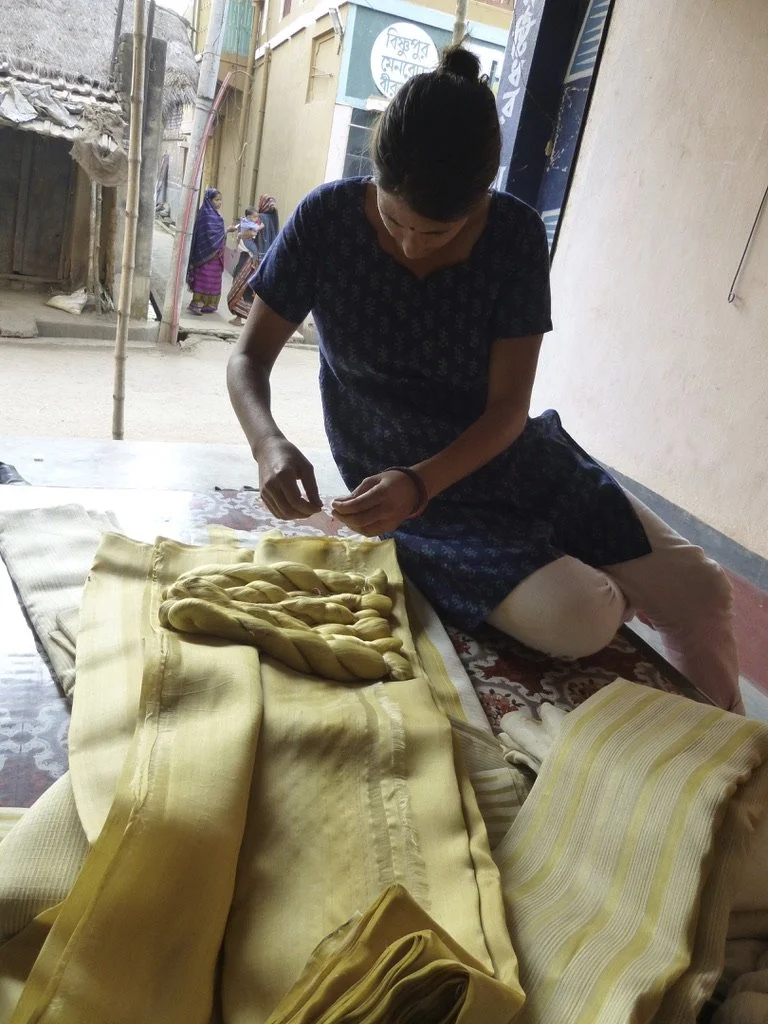 Woman sitting on floor weaving yellow fabric, surrounded by more folded fabrics, with outdoor scene and other people in background.