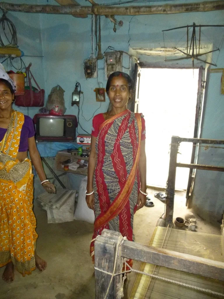 Two women in traditional sarees standing inside a small, modest room with a blue wall, cluttered with a small television, wires, and household items. One woman is smiling and facing forward, while the other is partially visible on the left.