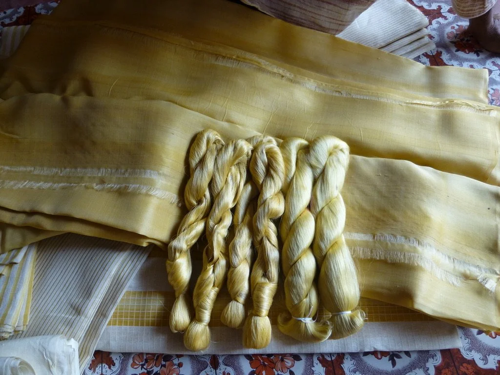 Several bundles of blond hair extensions or very long, blonde synthetic hair on top of a yellow silk fabric. The background shows patterned tablecloth and cloth napkins.