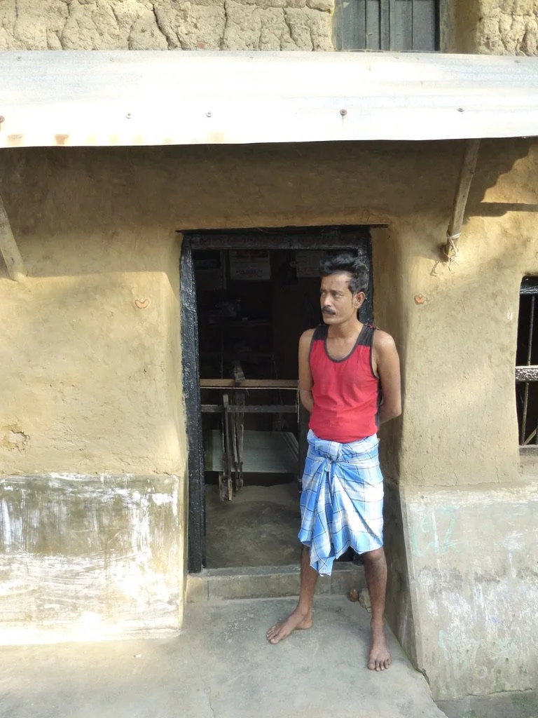 Man standing barefoot outside a small, rustic building with a yellowish mud wall and a black doorframe, wearing a red tank top and a plaid garment wrapped around his waist.