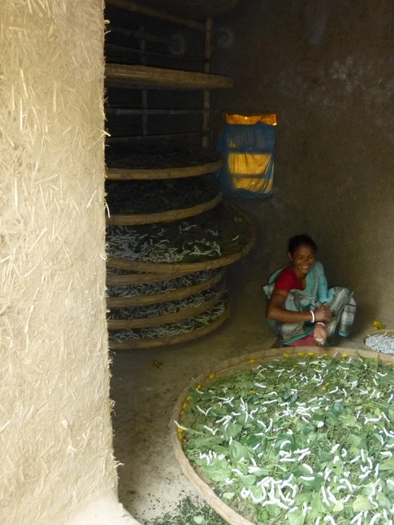 A woman inside a rustic room surrounded by large woven baskets holding green leaves, some with white and yellow flowers, and a smiling woman sitting on the ground near the baskets.