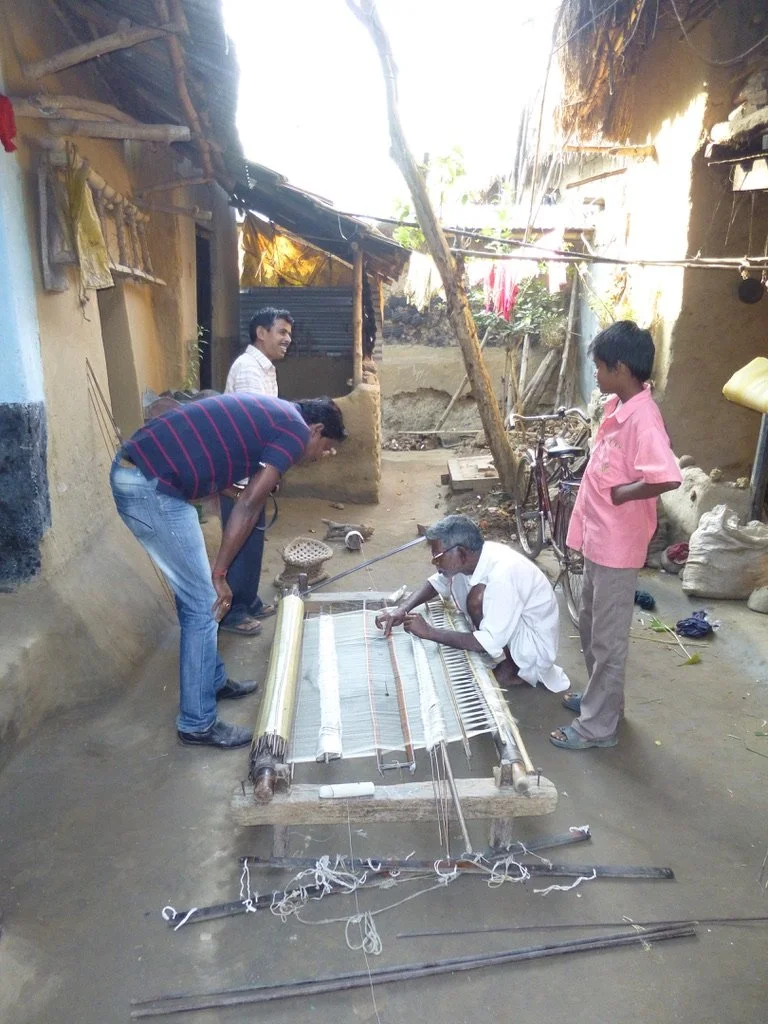 A man sits on the ground weaving a traditional loom, while four other people observe and engage with him outdoors in a rustic neighborhood.